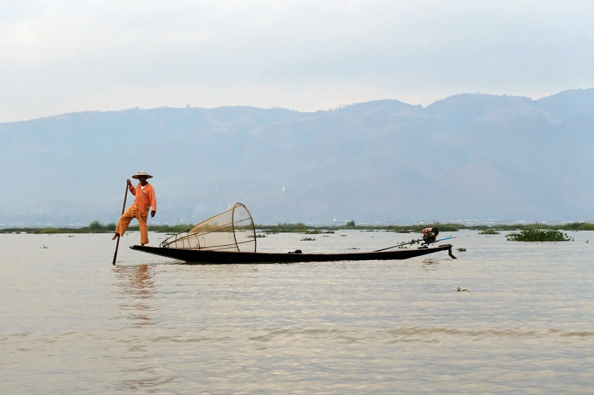 A person standing on a wooden boat with a fishing net in a lake, against a backdrop of hills. Myanmar