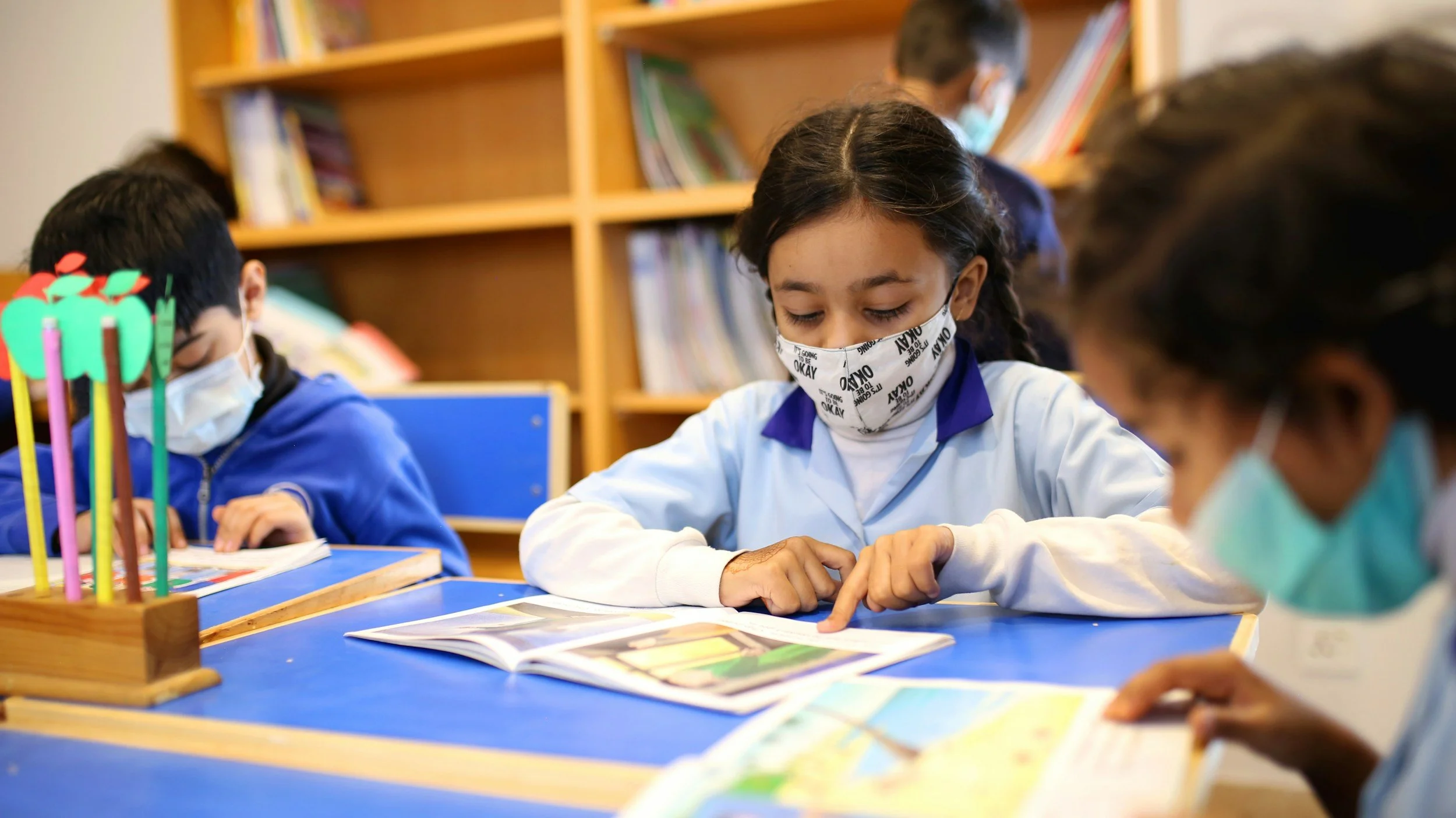 Students wearing masks sitting at desks in a classroom, looking at books.