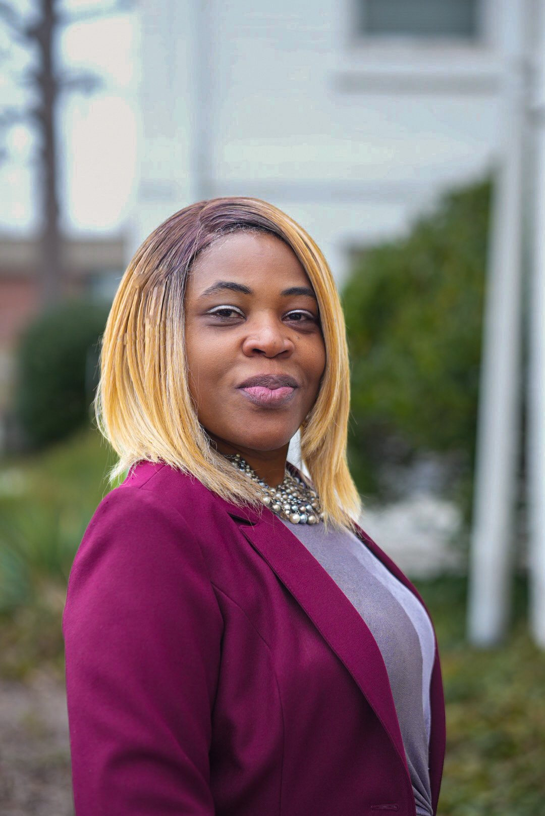 Portrait of a woman with shoulder-length blonde hair, wearing a purple blazer and a pearl necklace, standing outdoors with a blurred background of trees and a building.