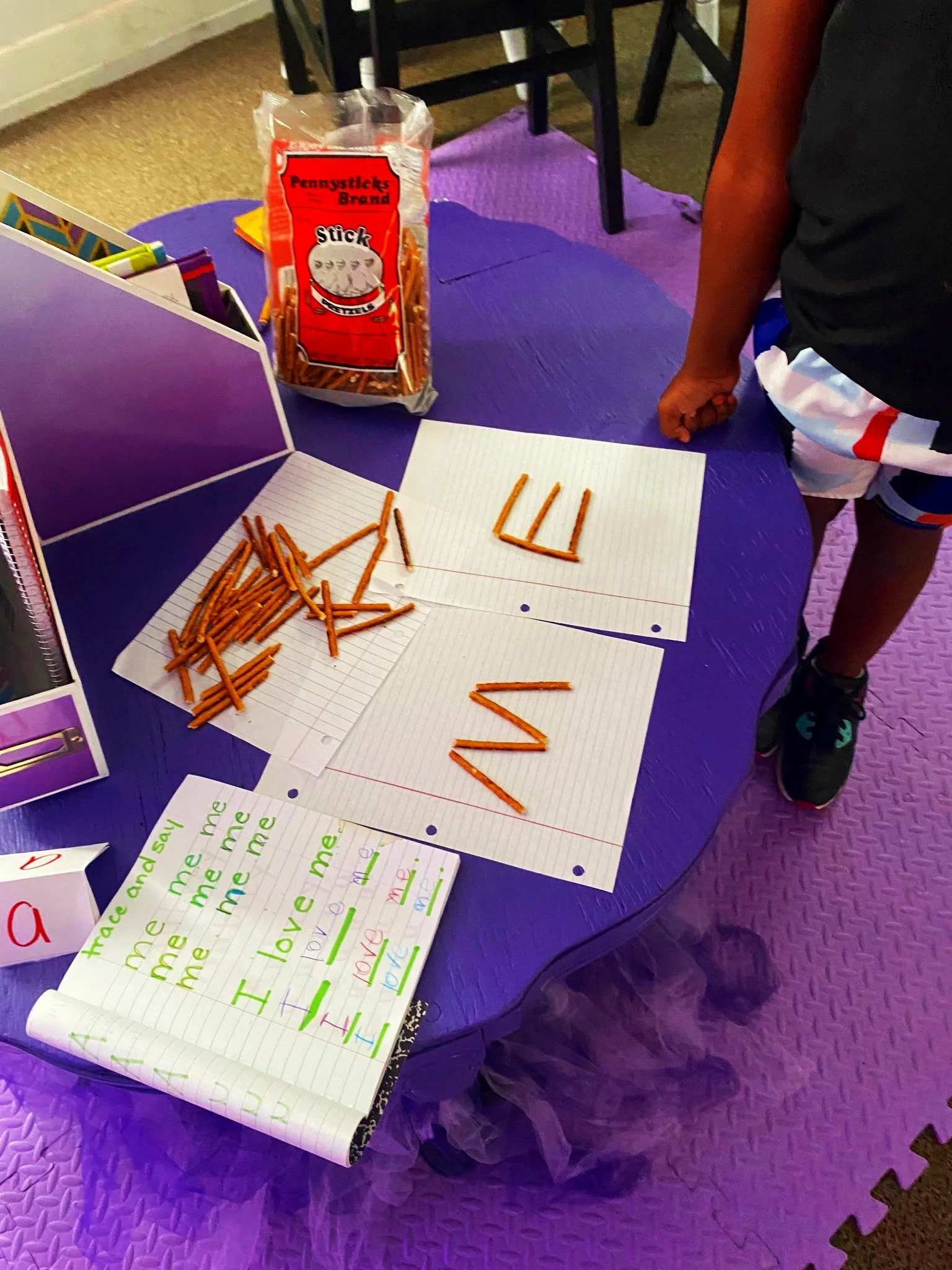 A purple table creating words with Stick pretzels, with letters made from pretzels, and handwritten notes about the word 'me' and the word 'love'.