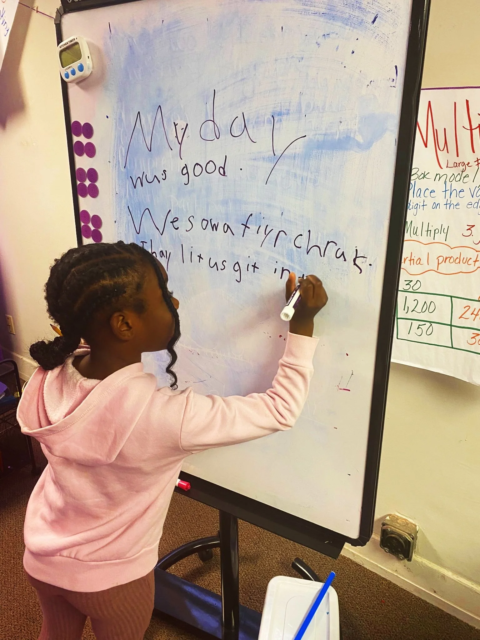 A young girl with braided hair, wearing a pink hoodie and brown pants, writes on a whiteboard with a dry-erase marker. The text she writes is: "Monday was good. We saw a fire truckt"