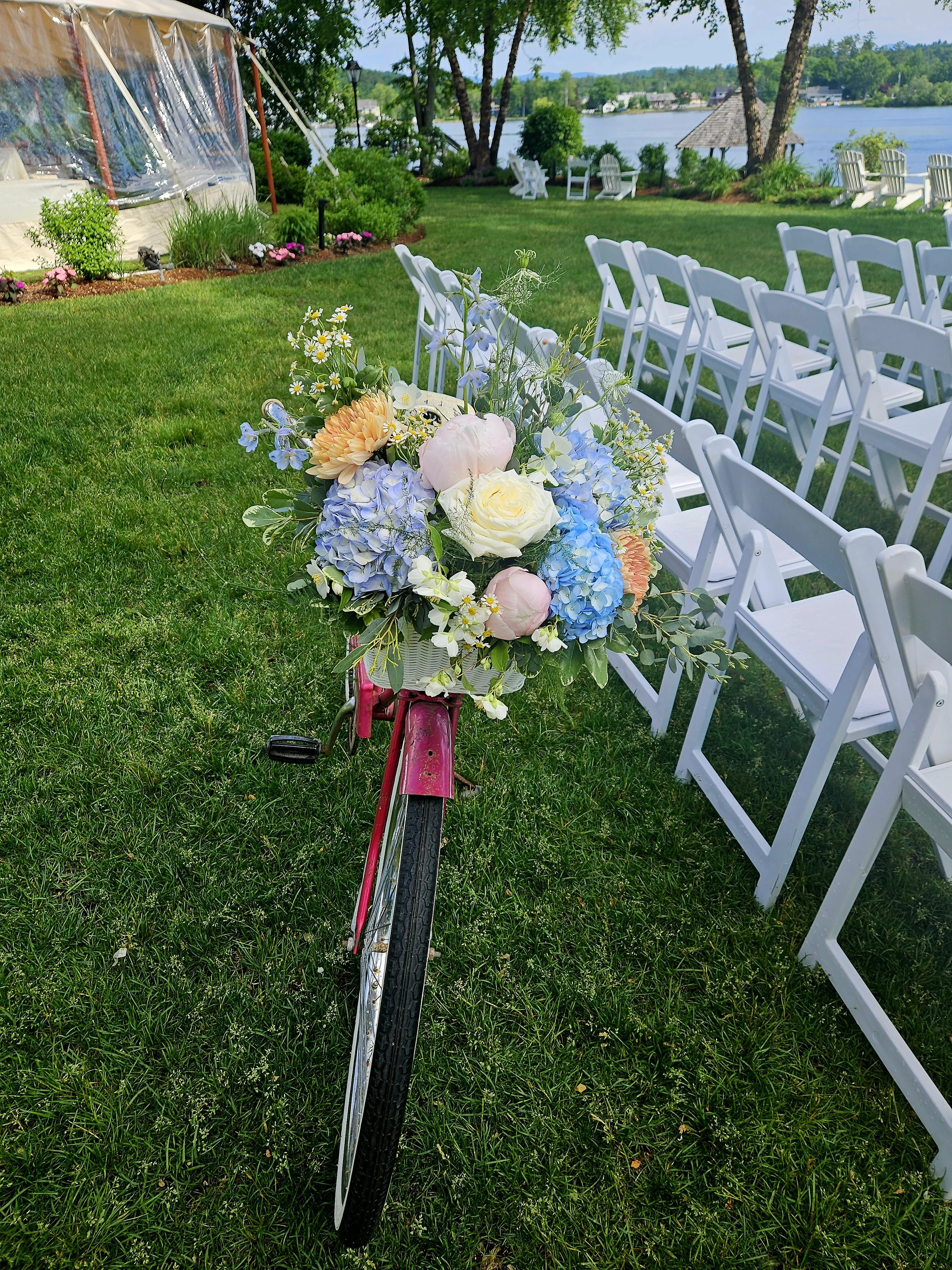 A row of white folding chairs arranged on a green lawn by a lake, with a bicycle decorated with a bouquet of pastel-colored flowers in the foreground.