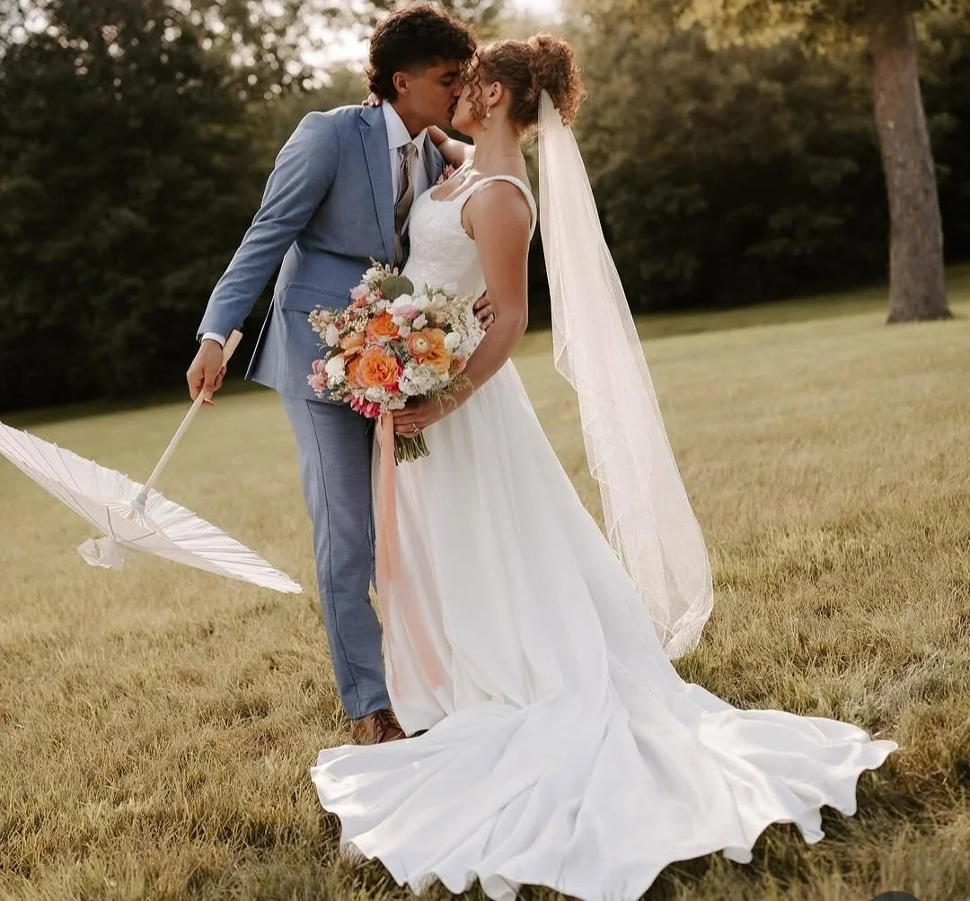 A bride and groom sharing a kiss outdoors on their wedding day. The bride is holding a bouquet of flowers and wearing a white wedding gown and veil, while the groom is dressed in a blue suit and holding a white umbrella.