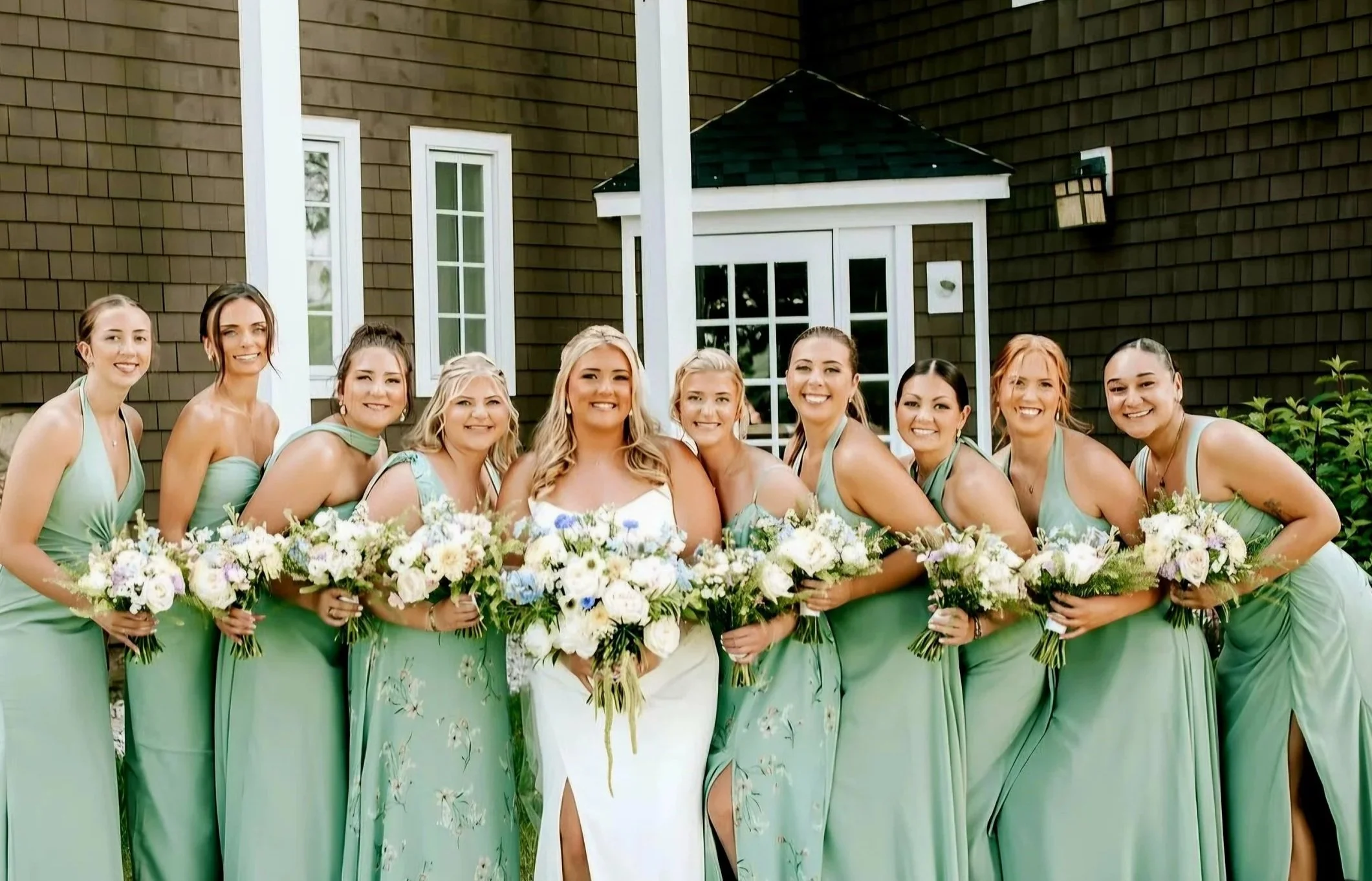 Group of women in matching teal dresses holding bouquets, smiling, standing outside a house with brown siding and white-framed windows.
