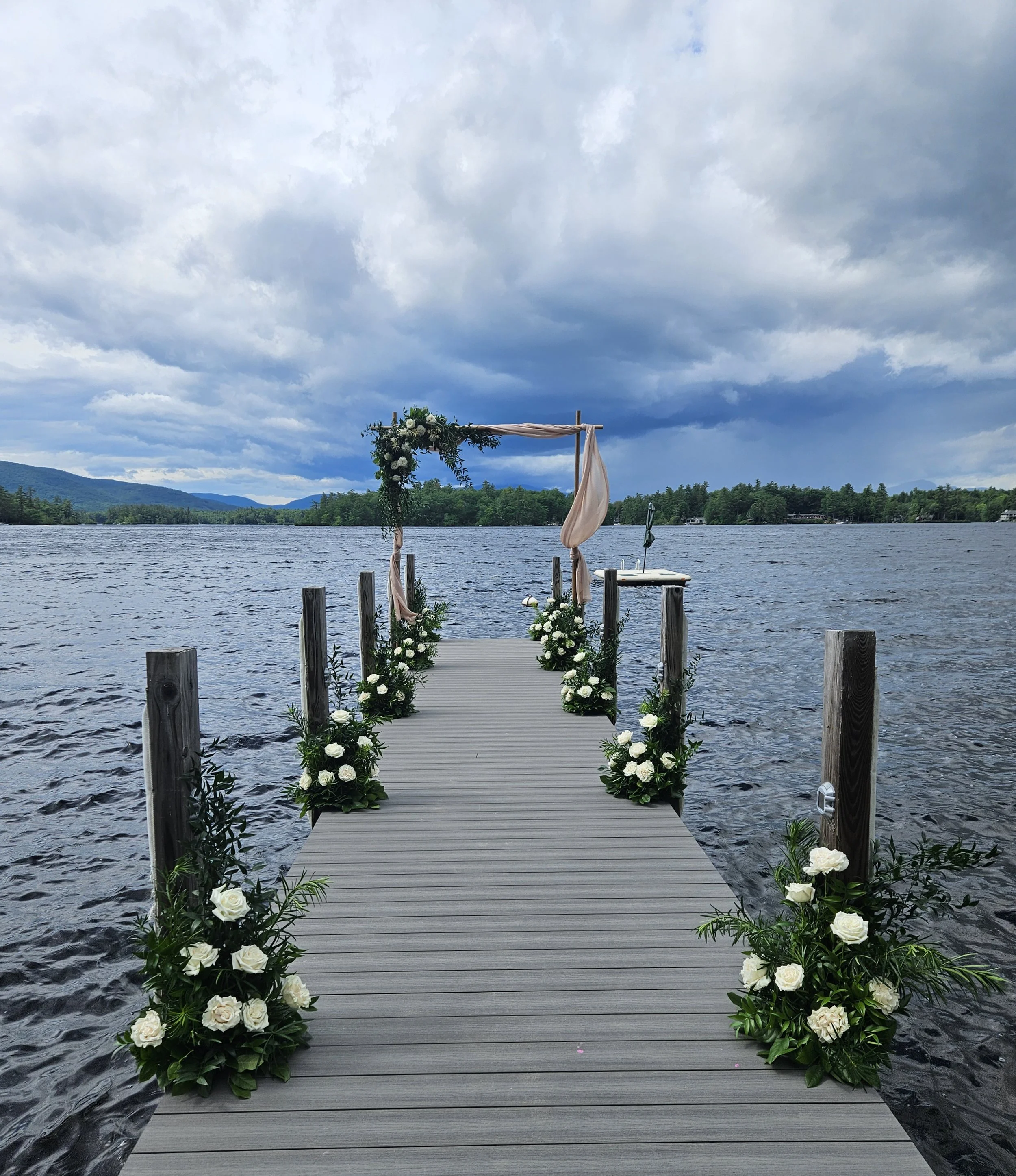 Wedding arch decorated with white flowers and greenery on a dock over a lake, with trees and mountains in the background under a cloudy sky.