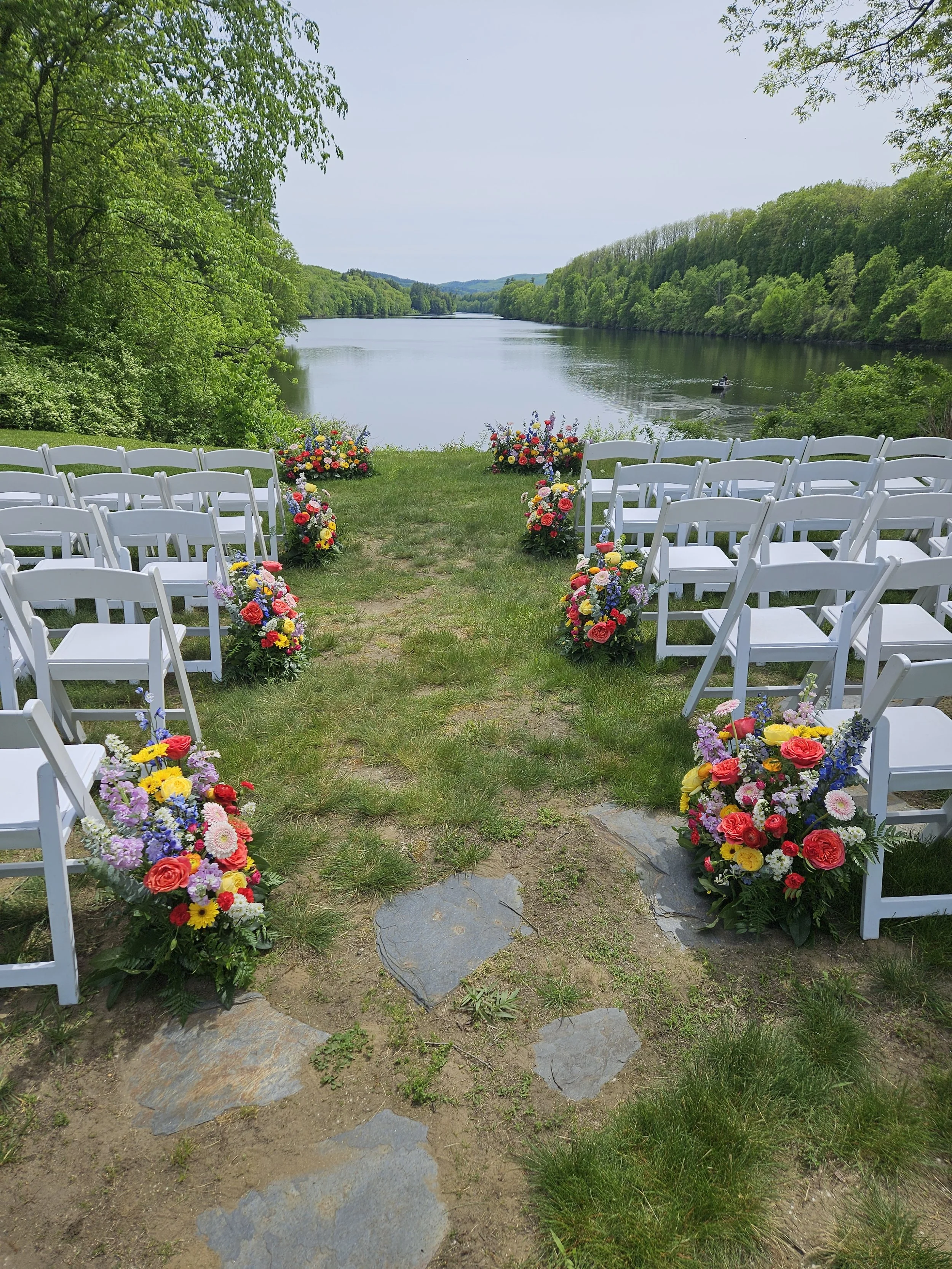 A lakeside outdoor wedding setup with white folding chairs decorated with colorful flower arrangements on each side of a grassy aisle, overlooking a calm lake surrounded by lush green trees and distant hills, under a clear blue sky.