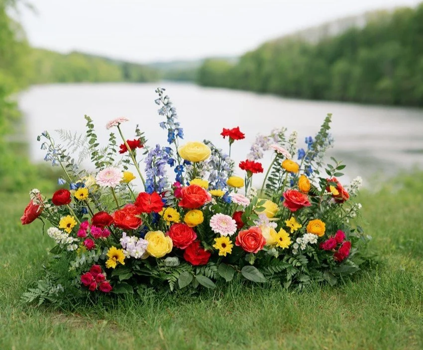 A vibrant bouquet of assorted colorful flowers placed on grass near a riverbank with green trees in the background.