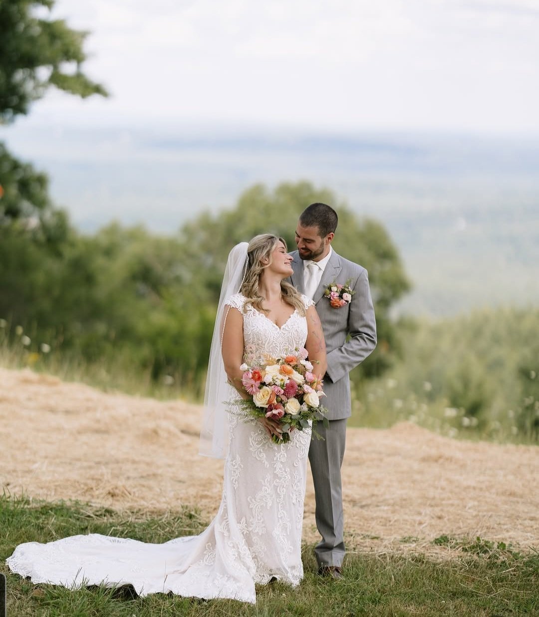 A bride and groom standing outdoors on their wedding day, sharing a moment and smiling at each other. The bride is in a white lace wedding dress holding a bouquet of pink, peach, and white flowers. The groom is in a light gray suit with a boutonniere matching the bouquet. The background features trees and a cloudy sky.