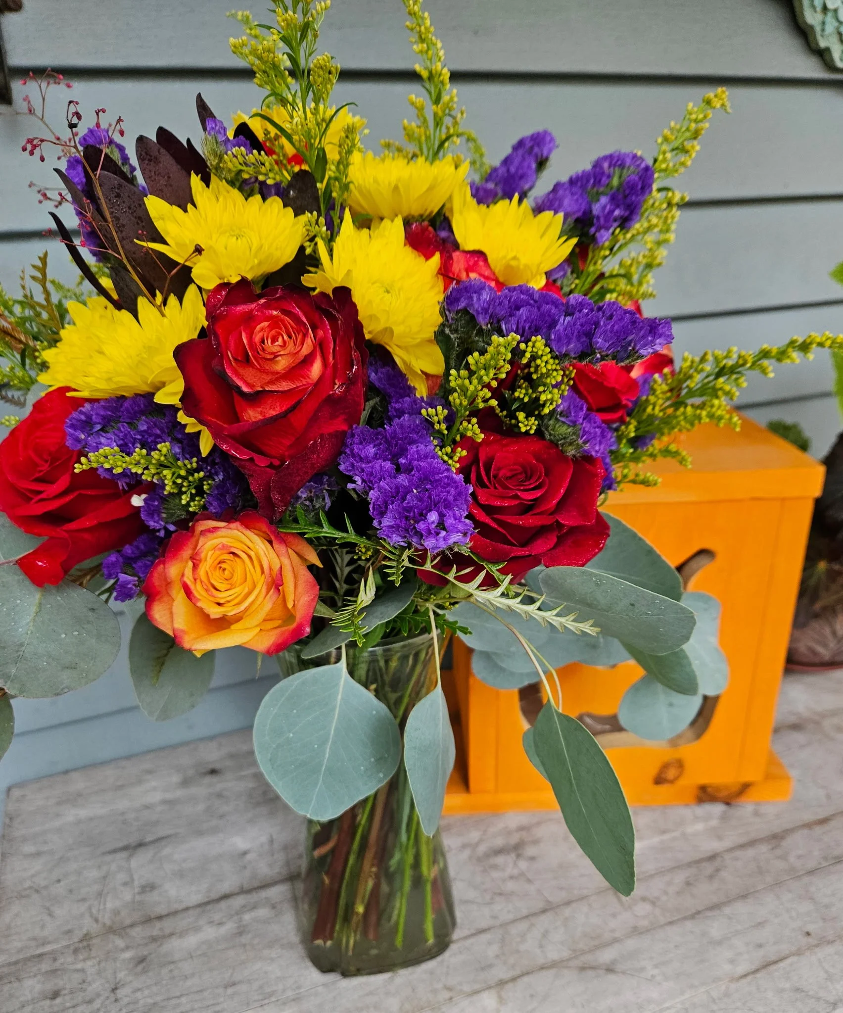 Bouquet of colorful flowers in a clear glass vase, placed on a gray wooden surface, with a wooden orange-colored box in the background.
