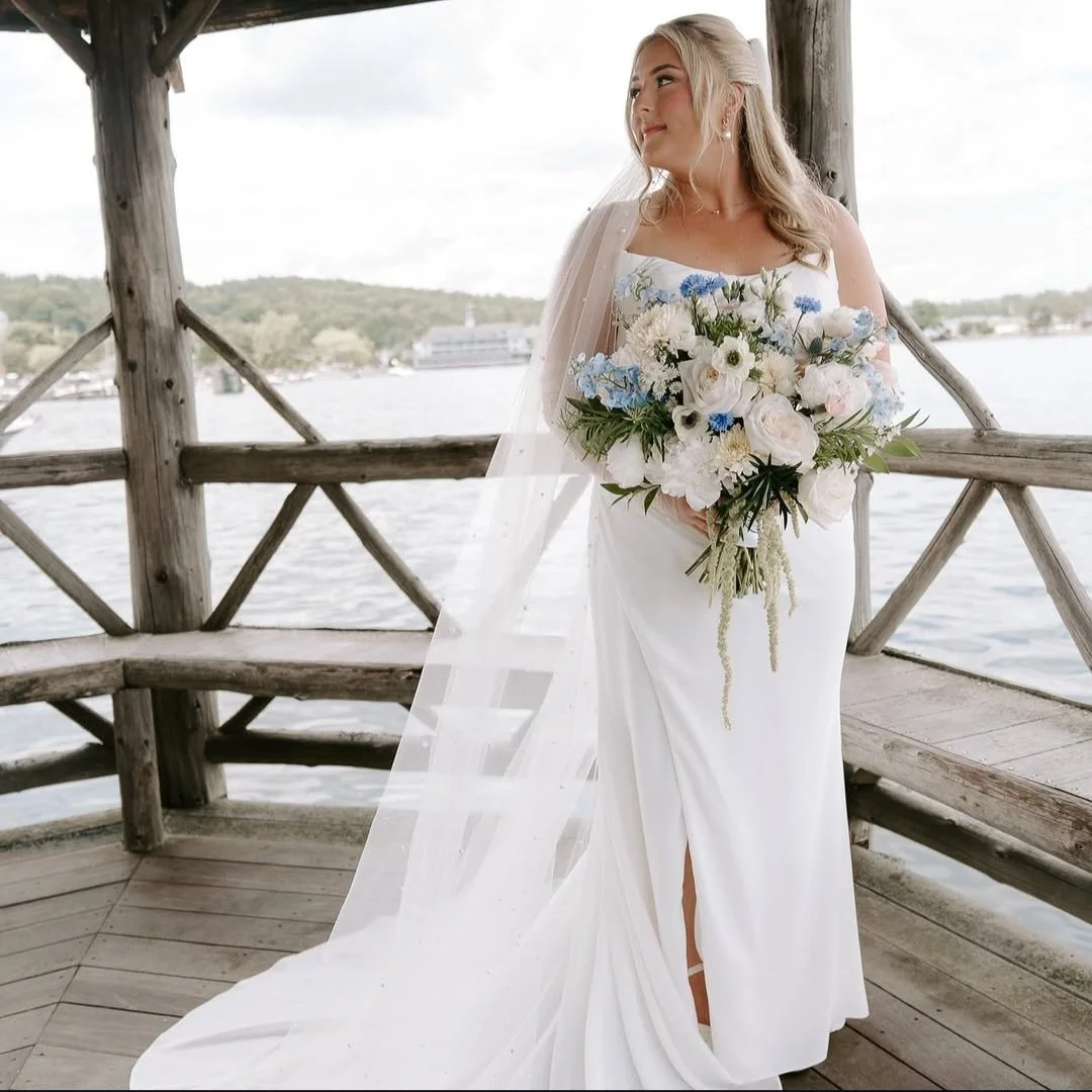 Bride in a white wedding dress holding a bouquet of white, blue, and pink flowers on a wooden dock by the water, with a veil and outdoor scenery in the background.