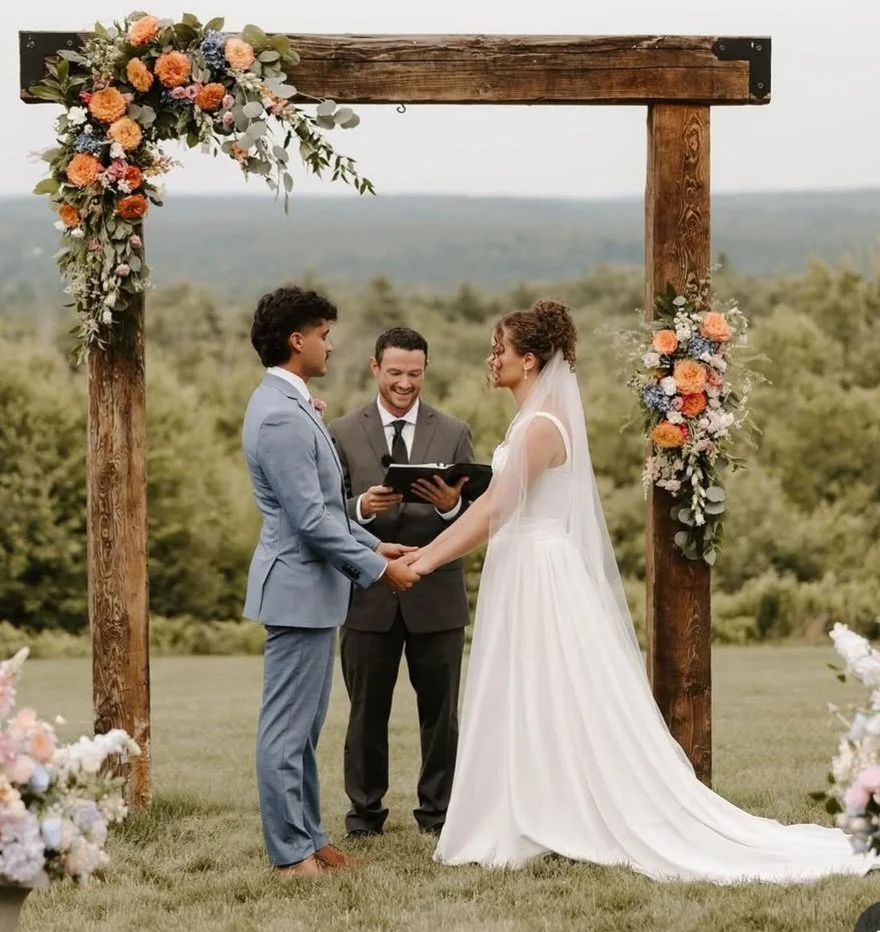 A couple getting married outdoors under a wooden arch decorated with flowers, holding hands, with an officiant officiating with a book, greenery and trees in the background.