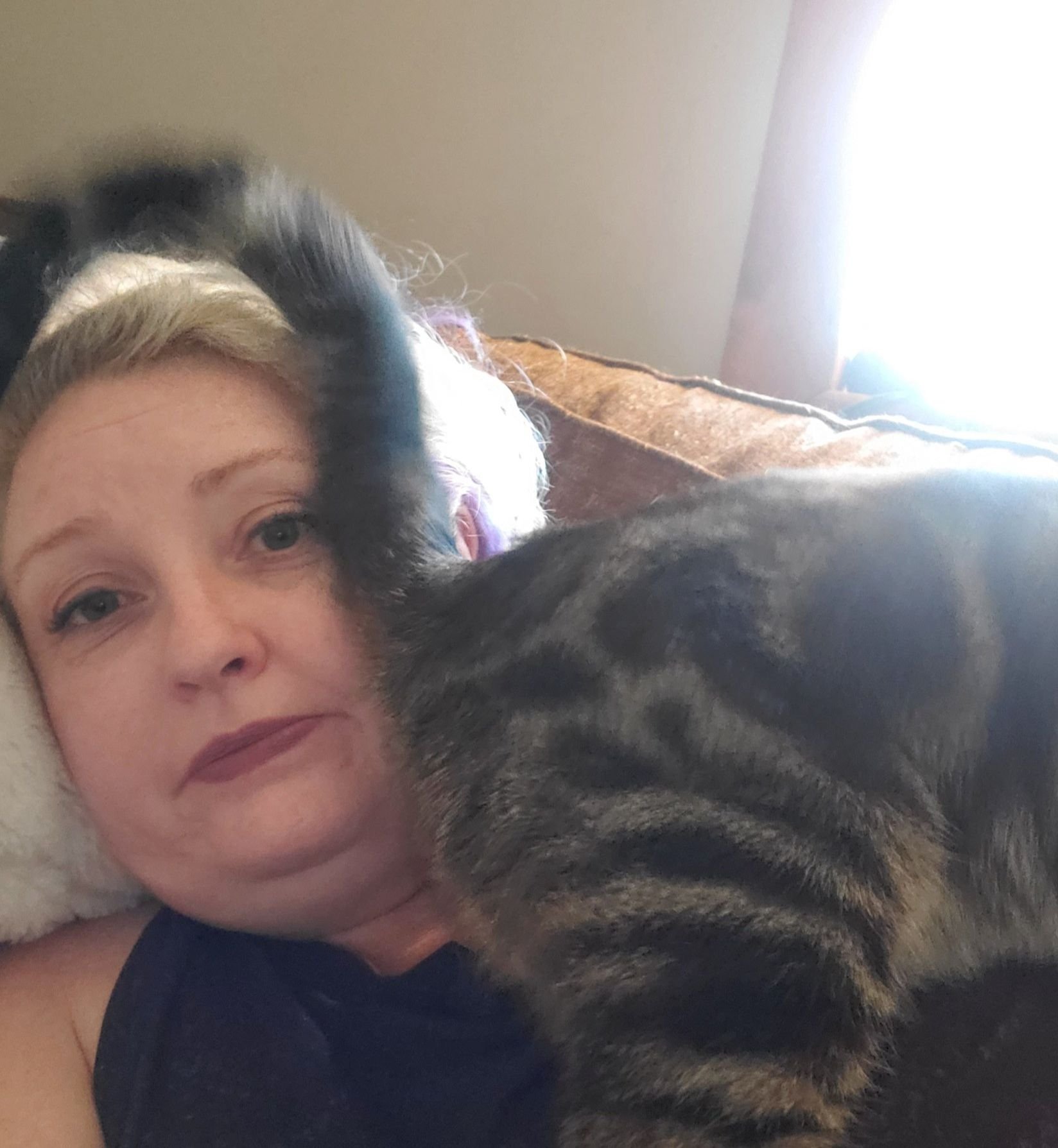 A woman lying on a pillow with a tabby cat resting her face. The woman has short blonde hair and is looking at the camera, while the cat is laying across her chest, partly obscuring her face.