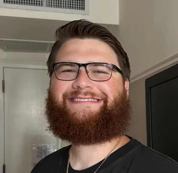 A young man with glasses and a beard smiling indoors.