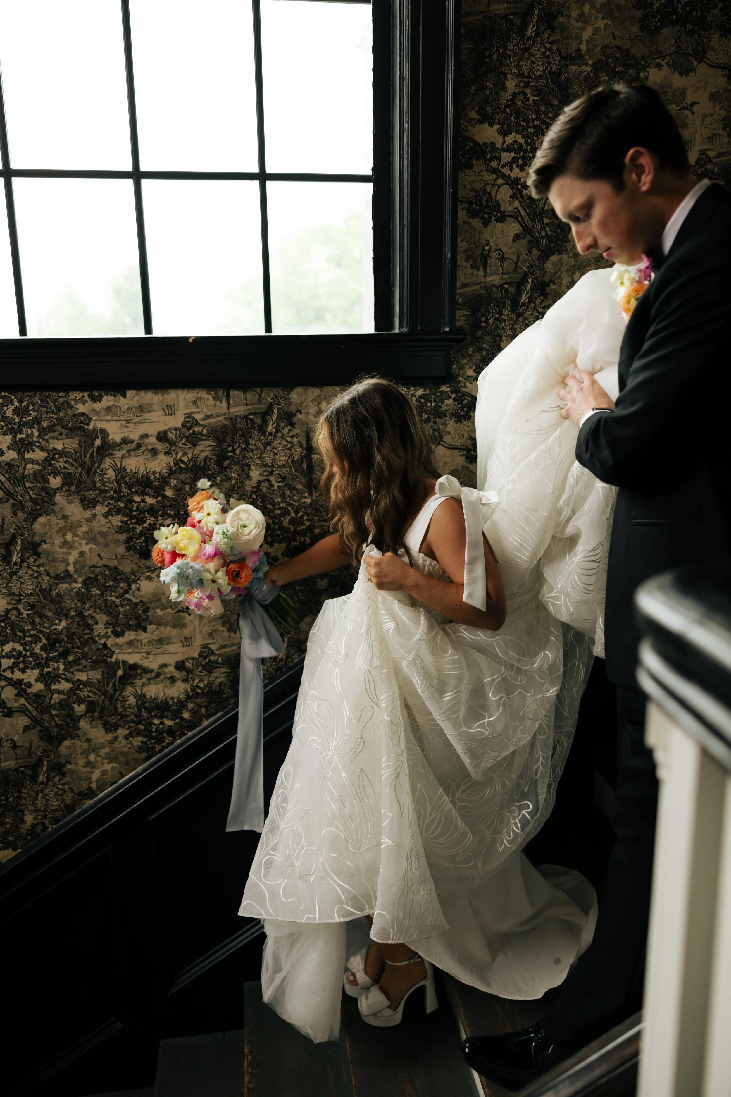 A bride in a white wedding dress, a young girl holding a bouquet, and a man in a dark suit, all standing on a staircase near a large window with black framing, inside a room with dark, patterned wallpaper.