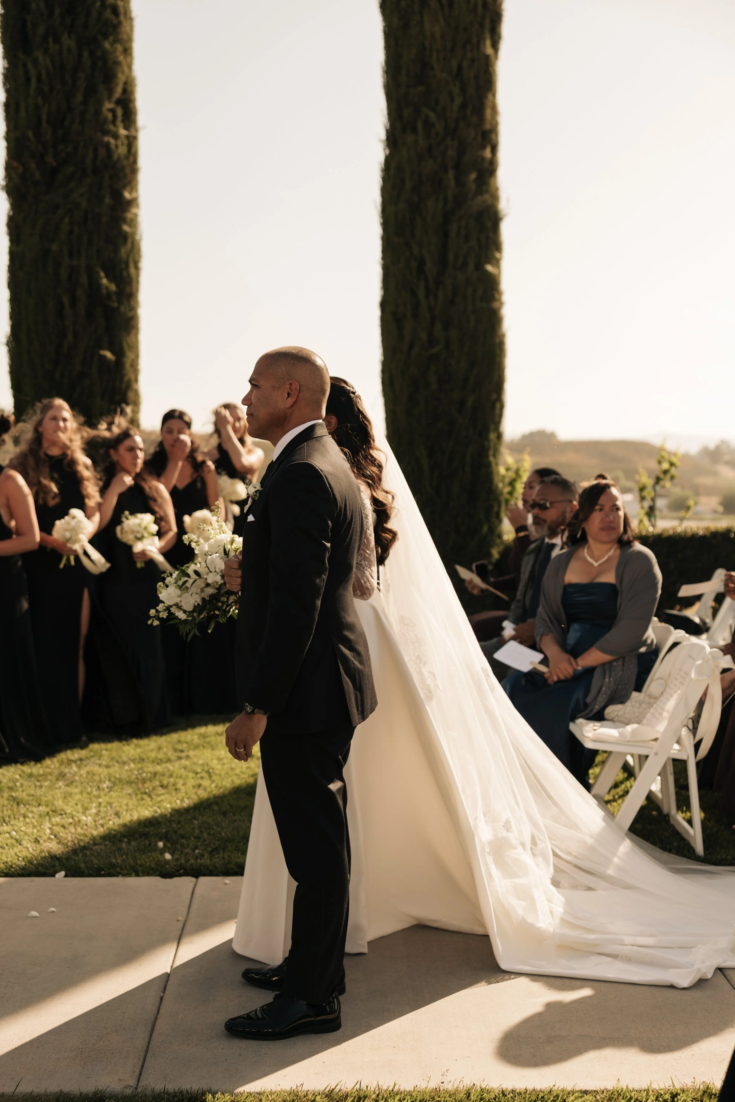 Groom standing during wedding ceremony outdoors with bridal party and guests in background. Temecula California wedding.
