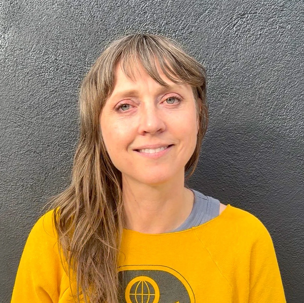 A woman with light brown hair and blue eyes smiling, wearing a yellow sweatshirt with a black globe logo, standing against a black textured wall.
