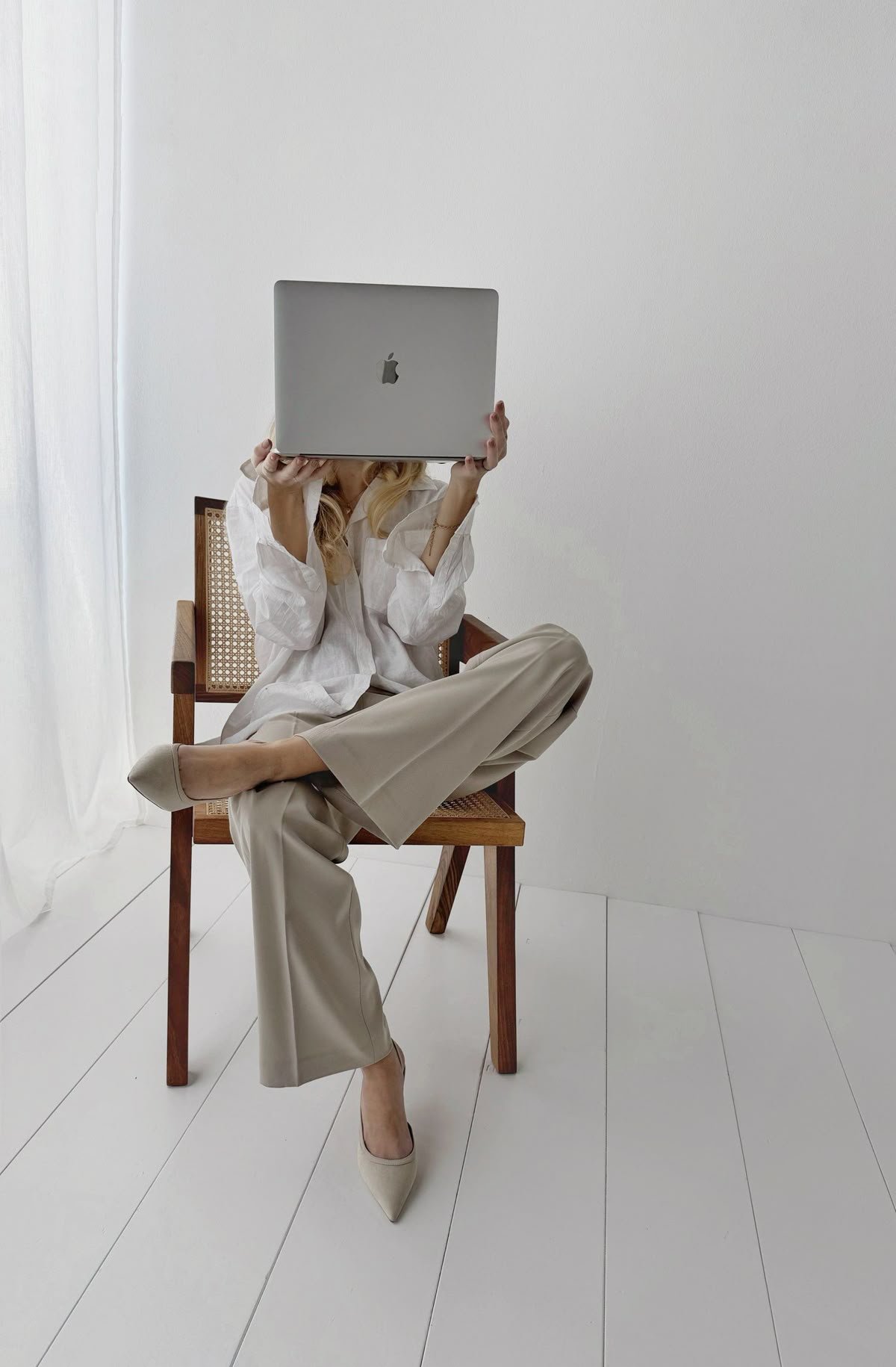A woman sitting cross-legged on a wooden chair, holding a silver MacBook computer in front of her face, with a plain white wall and white floor behind her.
