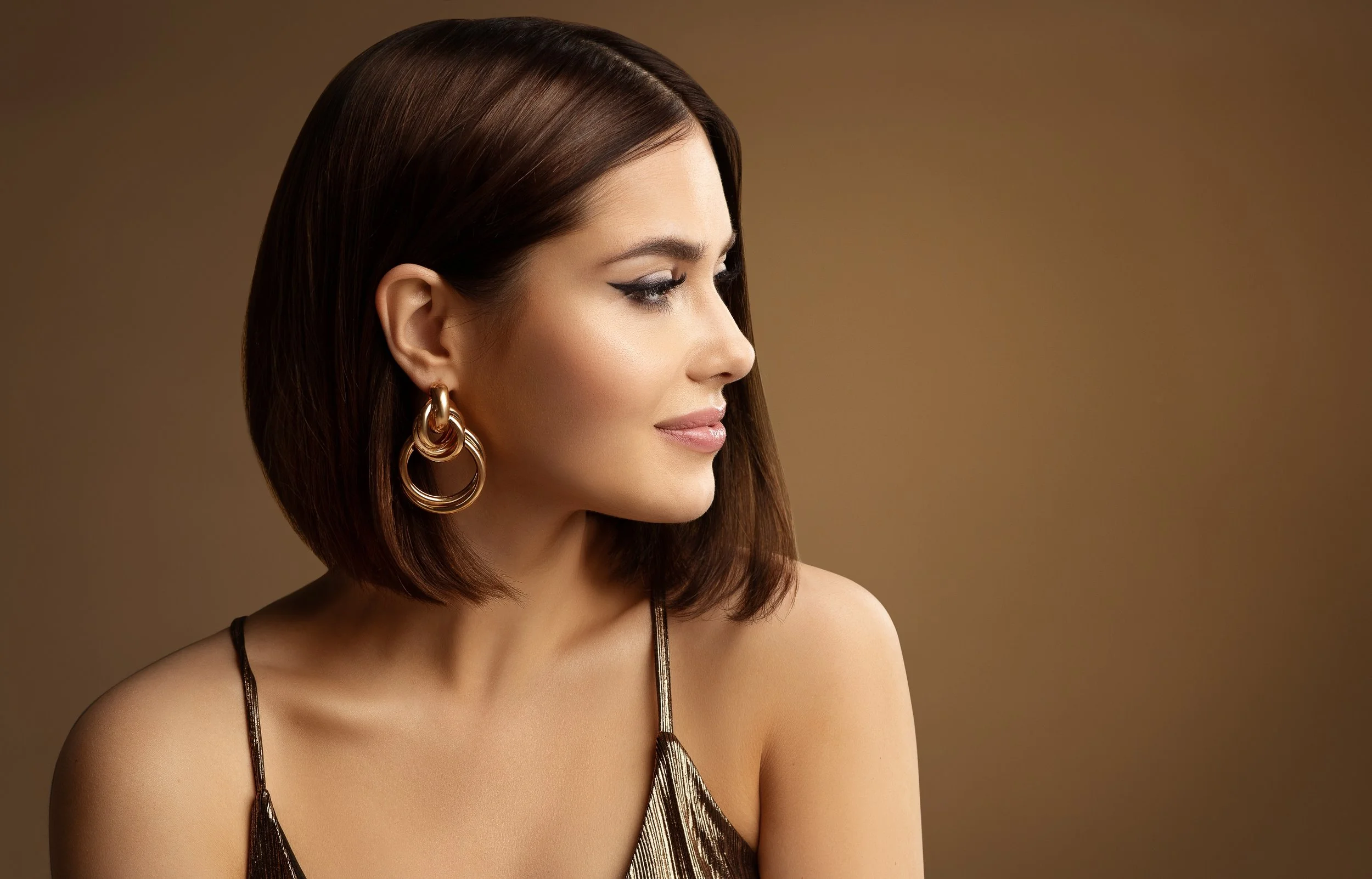 A woman with shoulder-length brown hair, wearing gold hoop earrings and a metallic dress, looking to her right against a plain brown background.