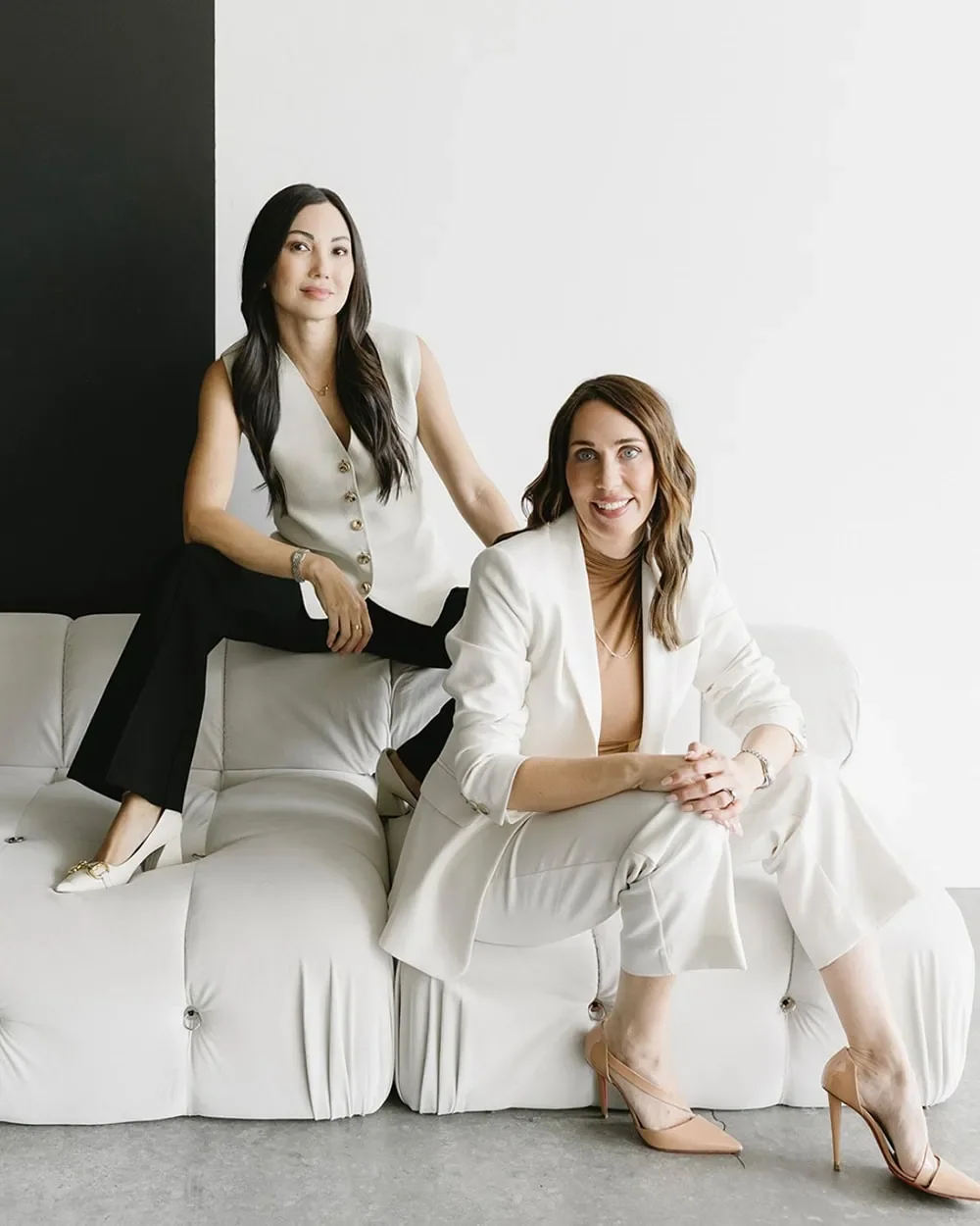 Two women in business attire sitting and standing on a white couch in a modern, minimalistic room, smiling and looking at the camera.