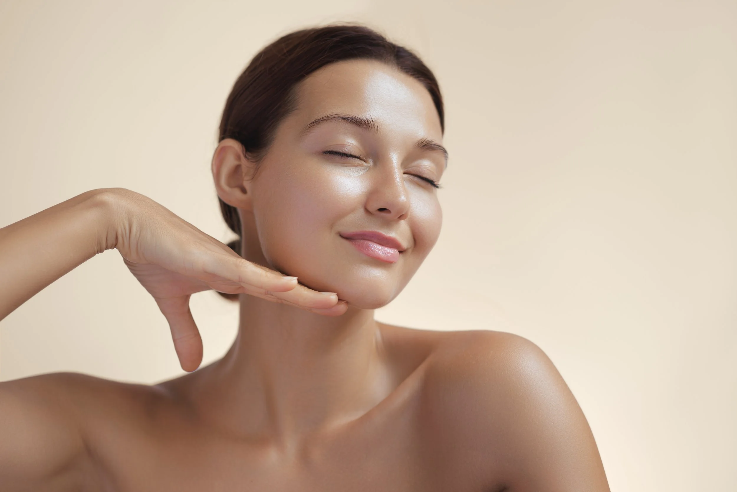 A woman with smooth, glowing skin gently resting her chin on her hand with a peaceful smile, against a plain beige background.