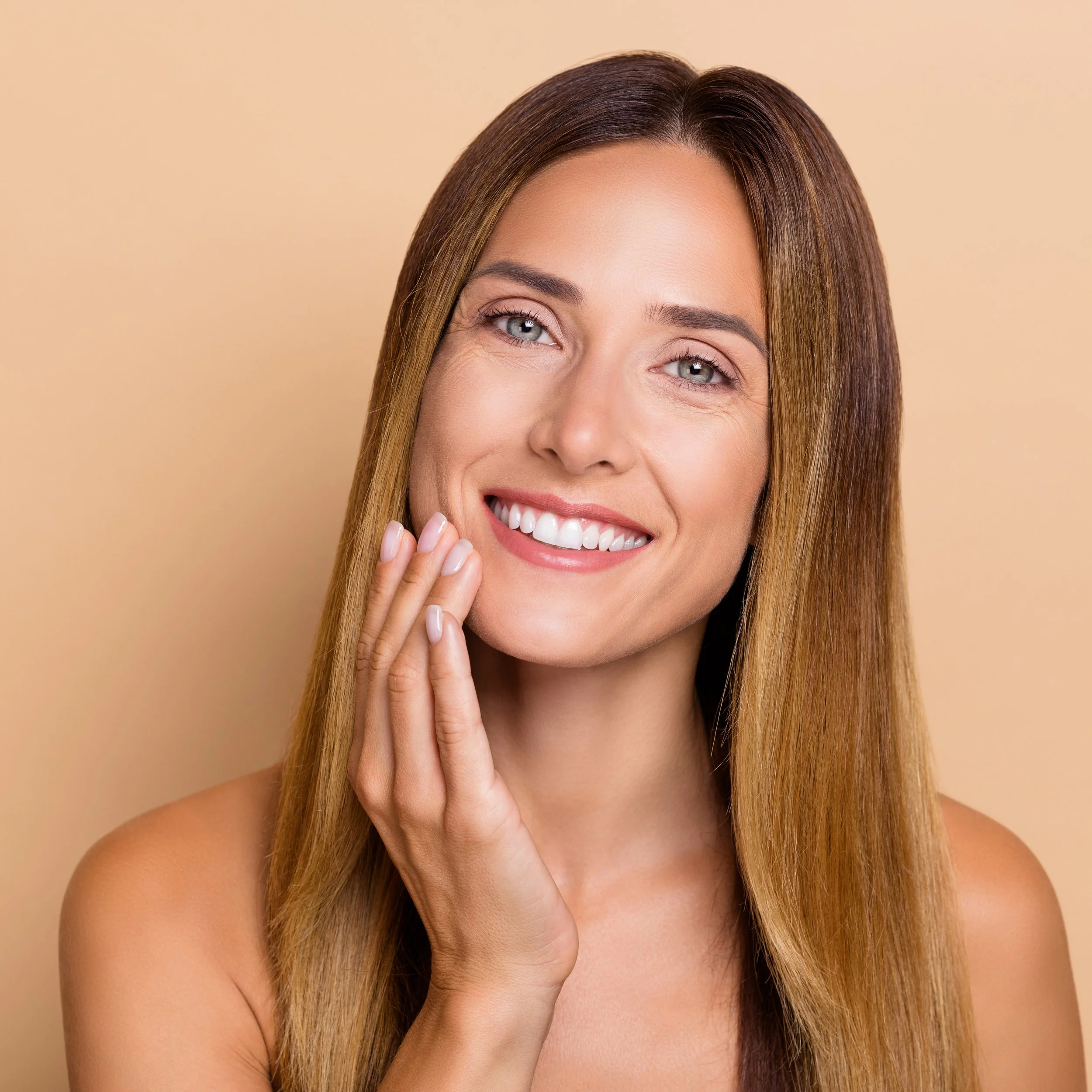 Close-up portrait of a smiling woman with long, straight brown hair, touching her face gently with her hand, against a beige background.