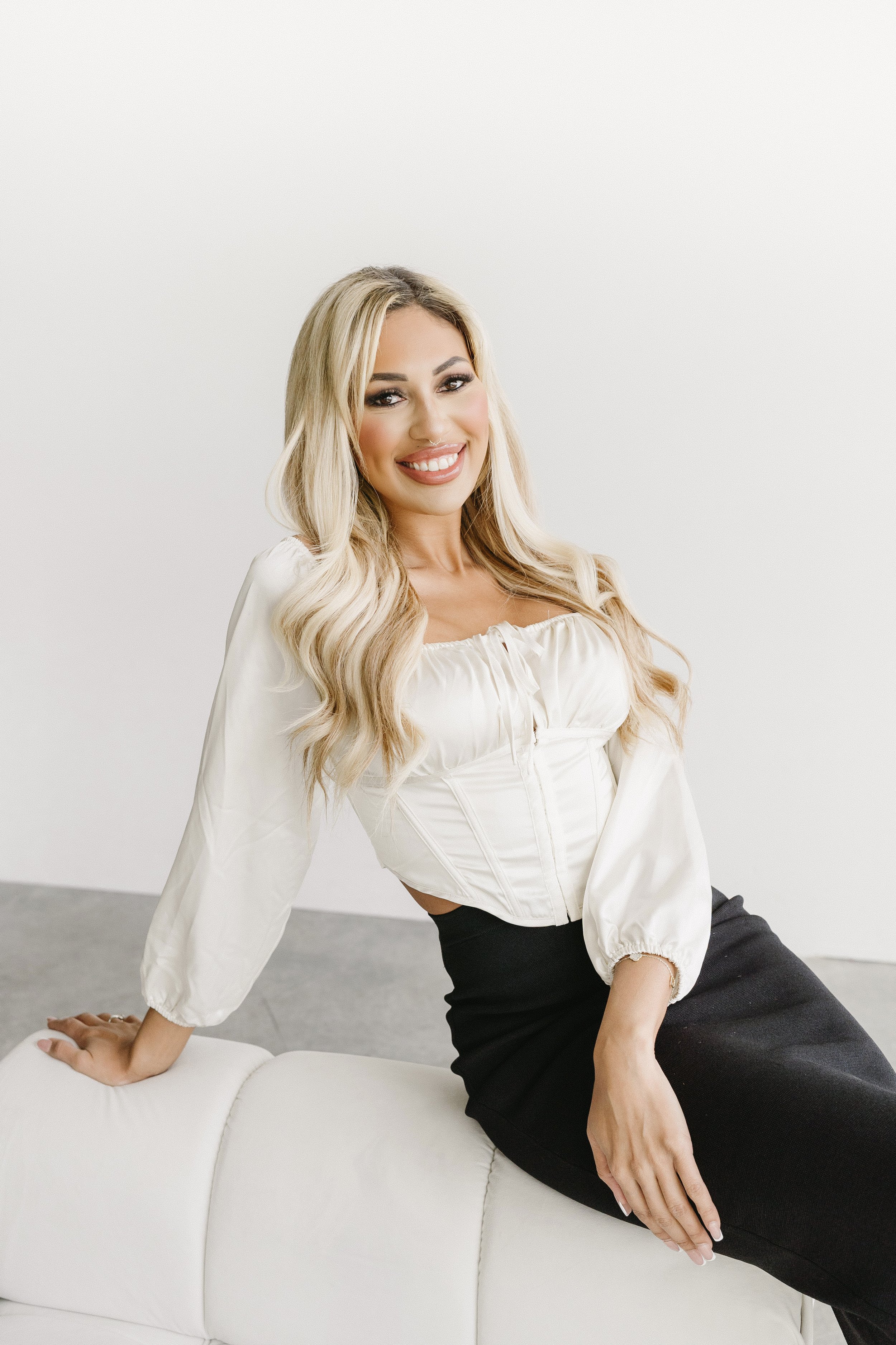 A woman with long blonde hair, wearing a white blouse and black skirt, sitting on a white couch, smiling.