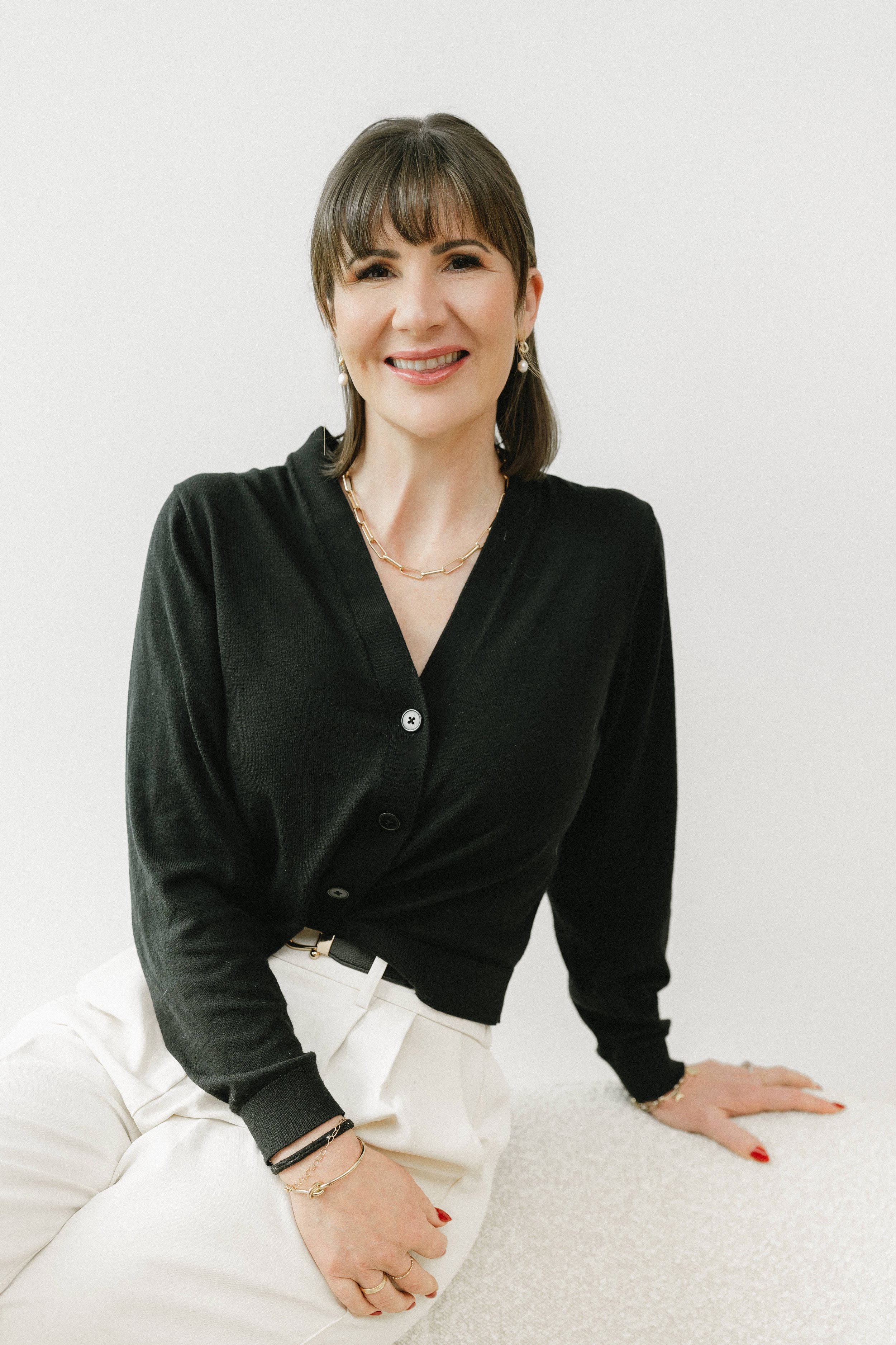 A woman smiling, wearing a black cardigan, white pants, and jewelry, sitting on a light-colored surface against a white background.