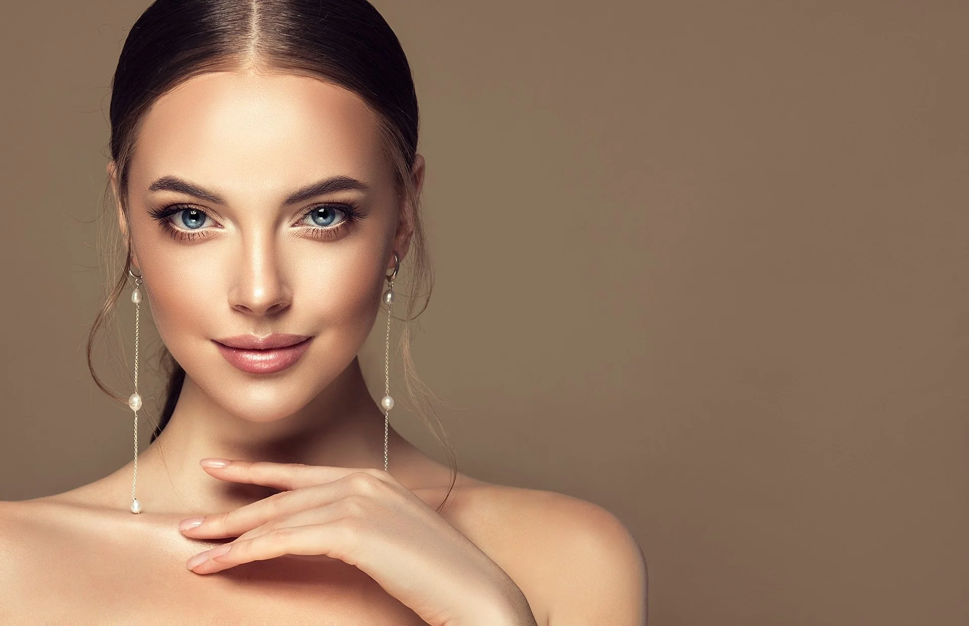 Close-up of a woman with fair skin, blonde hair tied back, blue eyes, wearing pearl drop earrings, with a subtle smile, against a plain beige background.