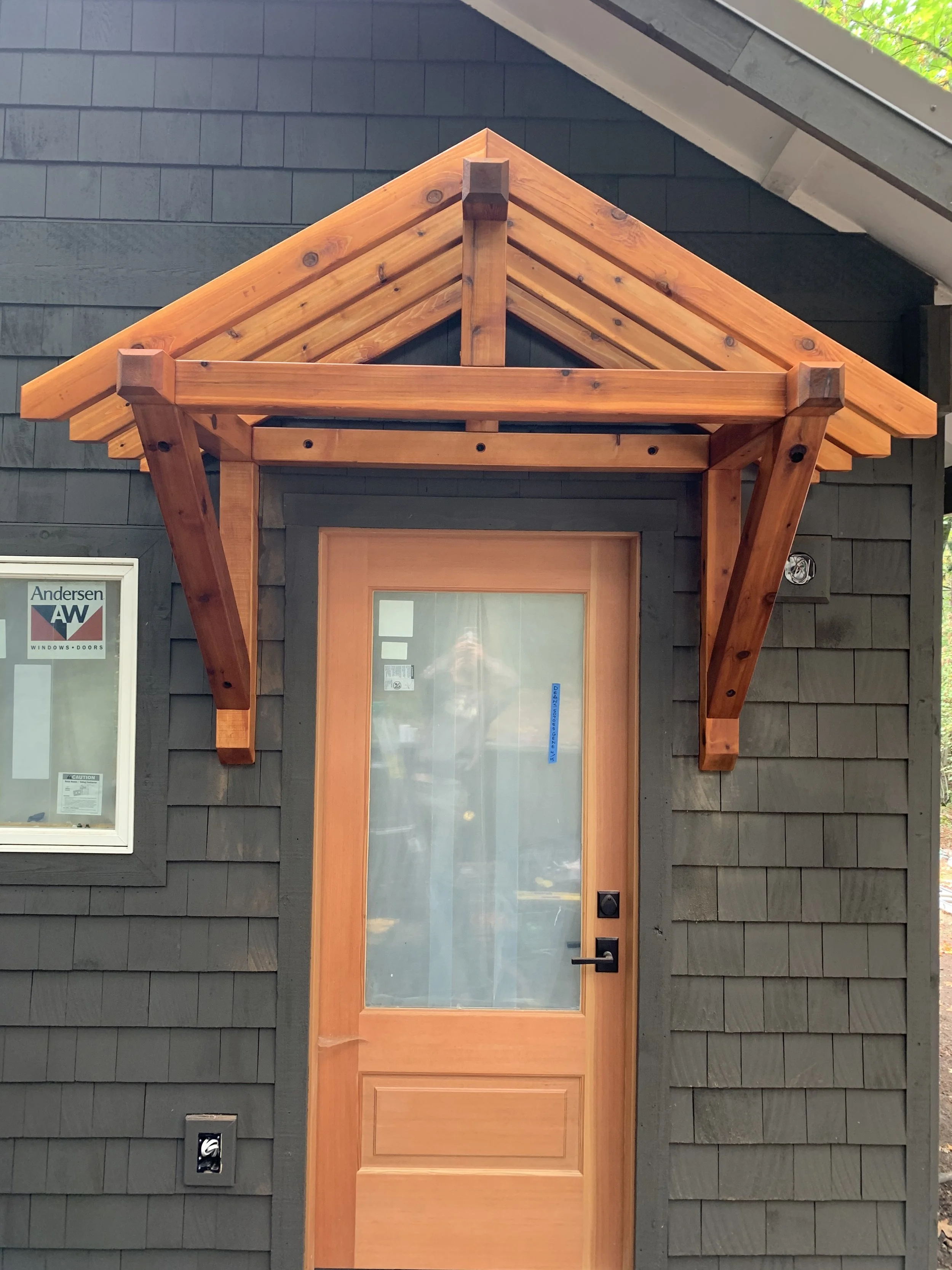 A wooden door with glass panel set in a black shingled exterior wall, with a small covered awning above it.