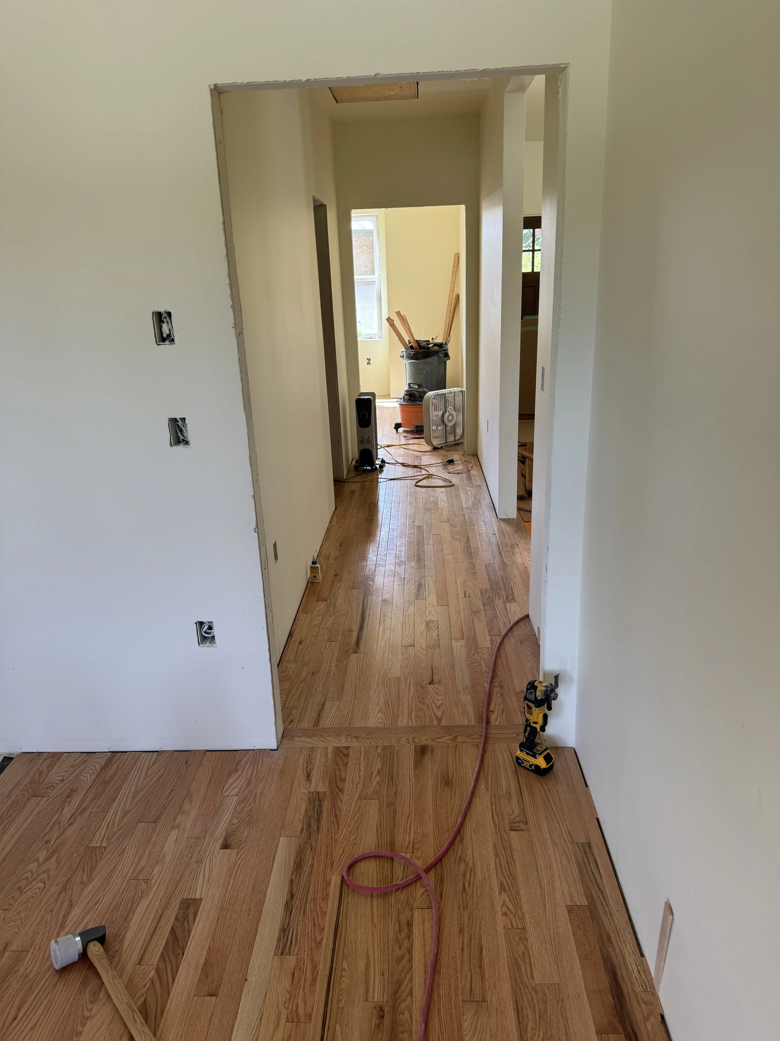 Interior view of a house under renovation, showing unfinished doorway, hardwood floor, construction tools, and supplies in the background.