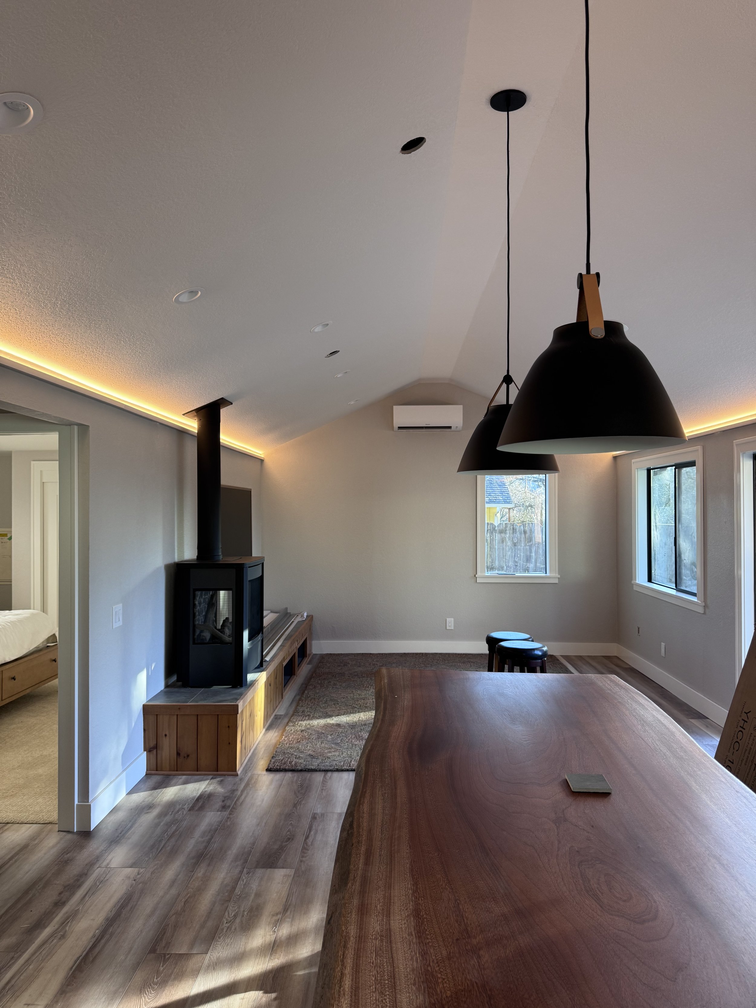 Living room with wooden dining table, hanging black pendant lights, windows, and a wood stove in the corner.