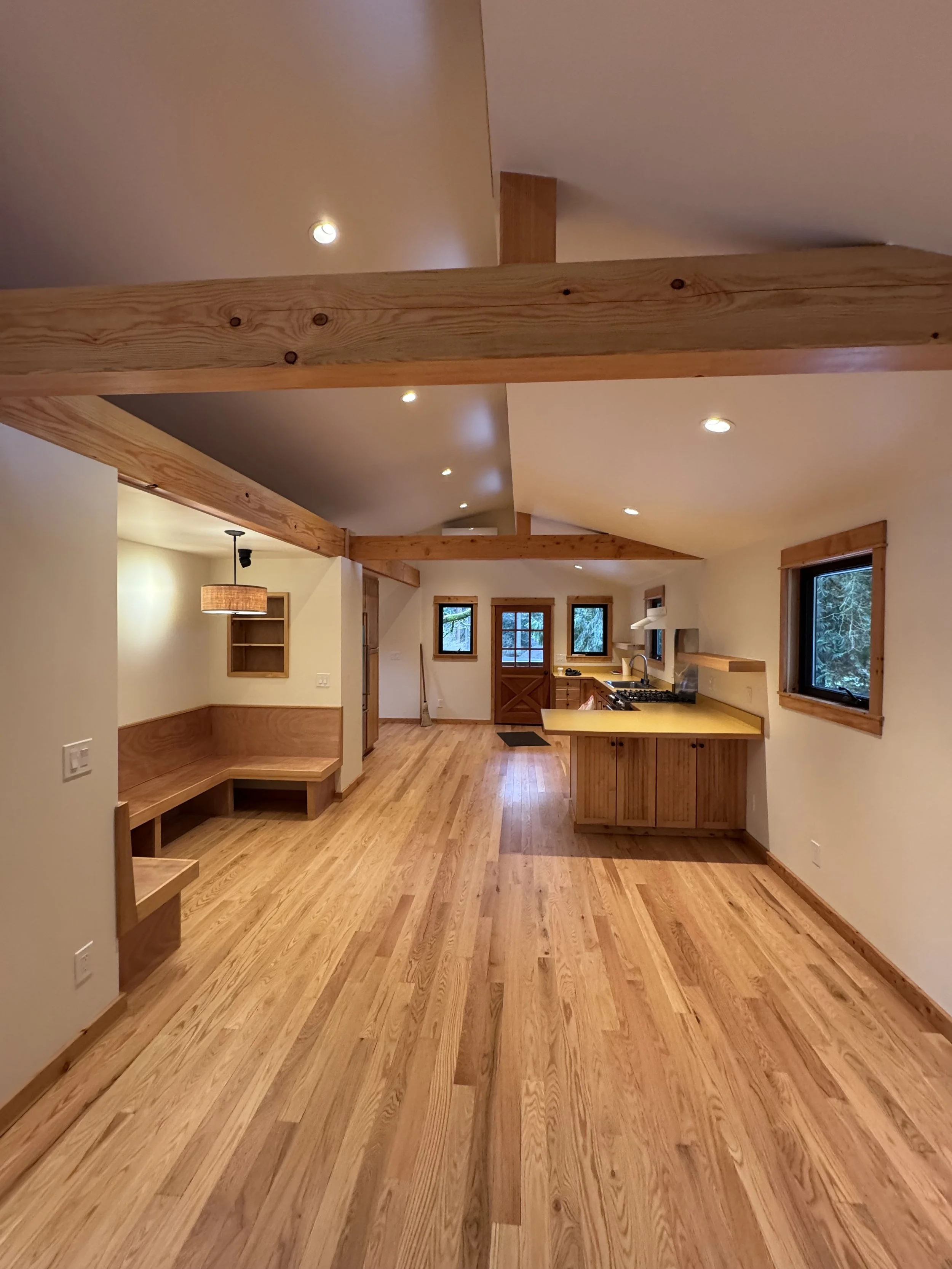 Interior view of a spacious room with wooden beams, hardwood floors, and multiple windows, featuring a kitchen area with a countertop, stove, and sink.