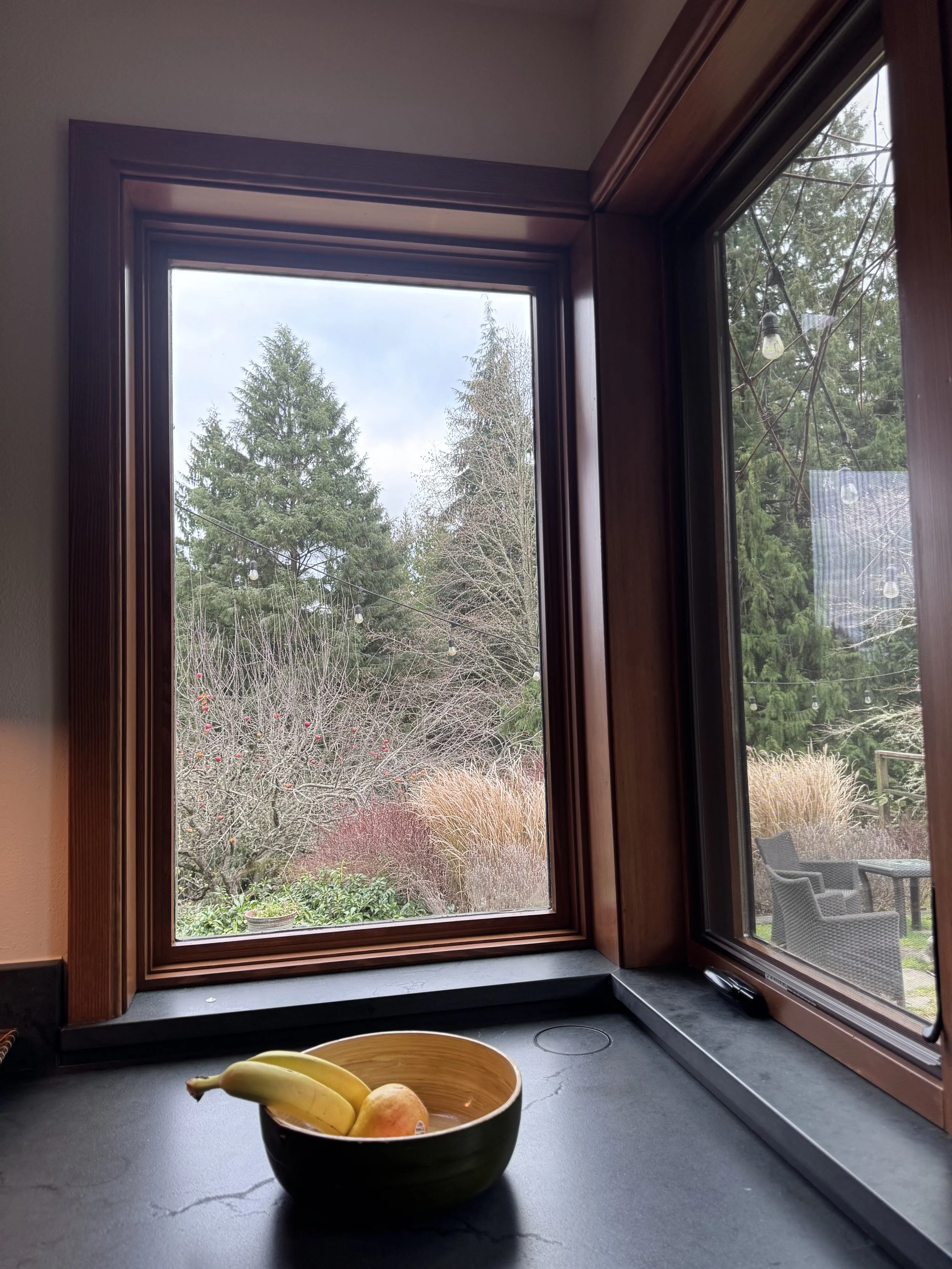 View from kitchen window showing trees, shrubs, and patio furniture outside with stone counters and vertical grain fir trim