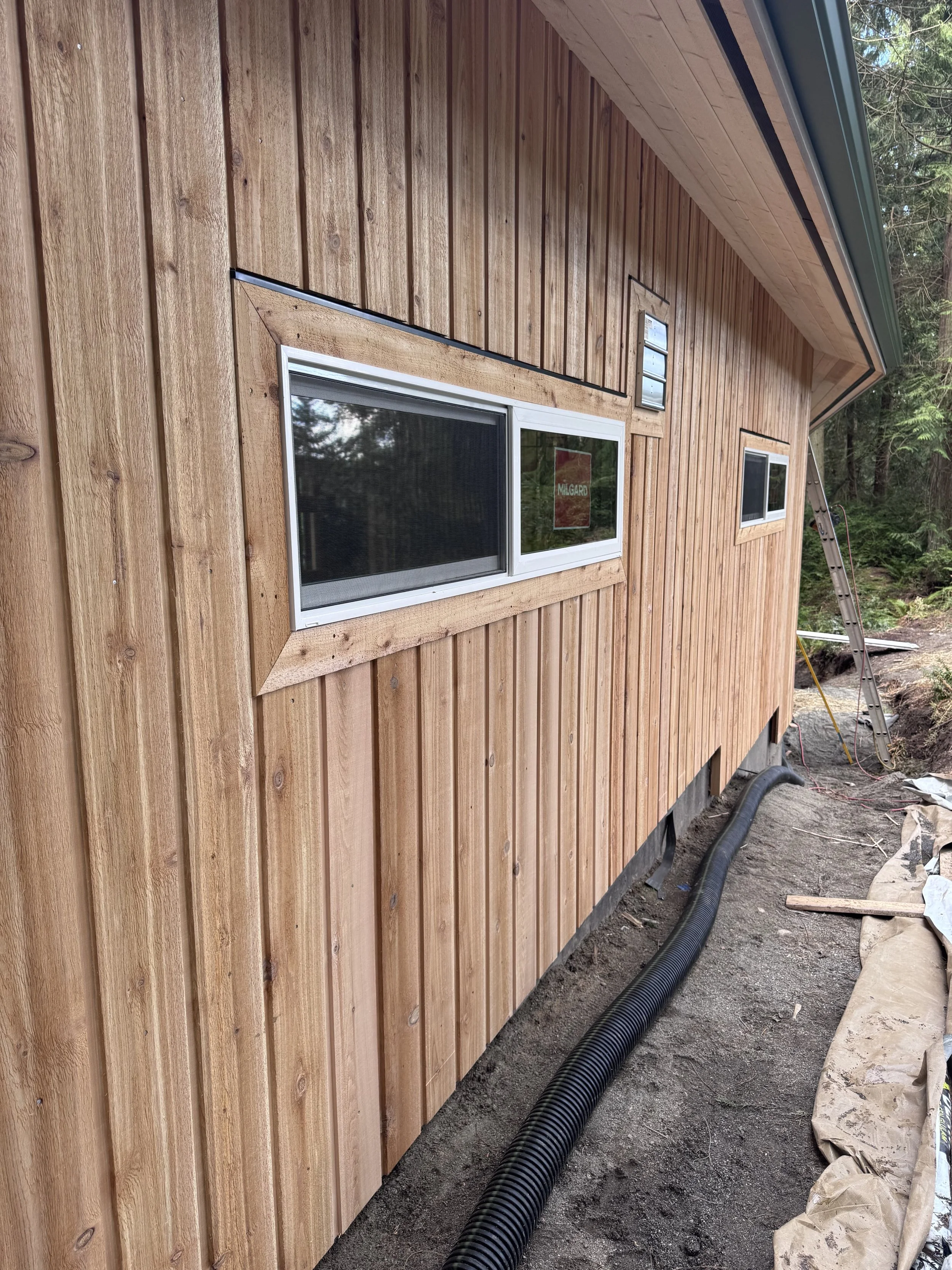 A wooden house exterior under construction, showing vertical wooden siding, rectangular windows, and black drainage pipes along the ground and forest in the background.