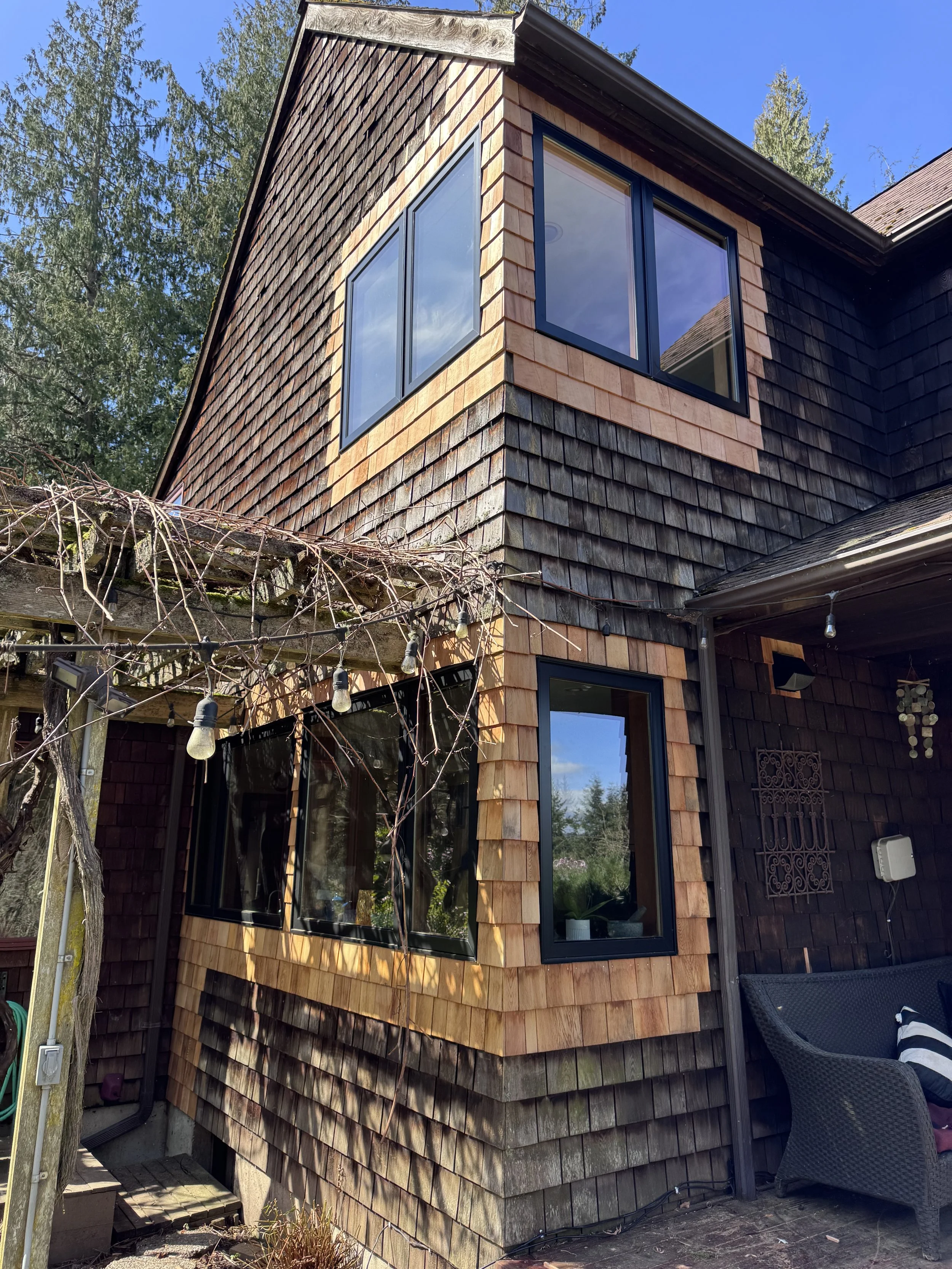 Close-up view of a multi-story house exterior with new wood shingles tied into the old shingles, large black-framed windows, a patio area, and a blue sky with trees in the background.