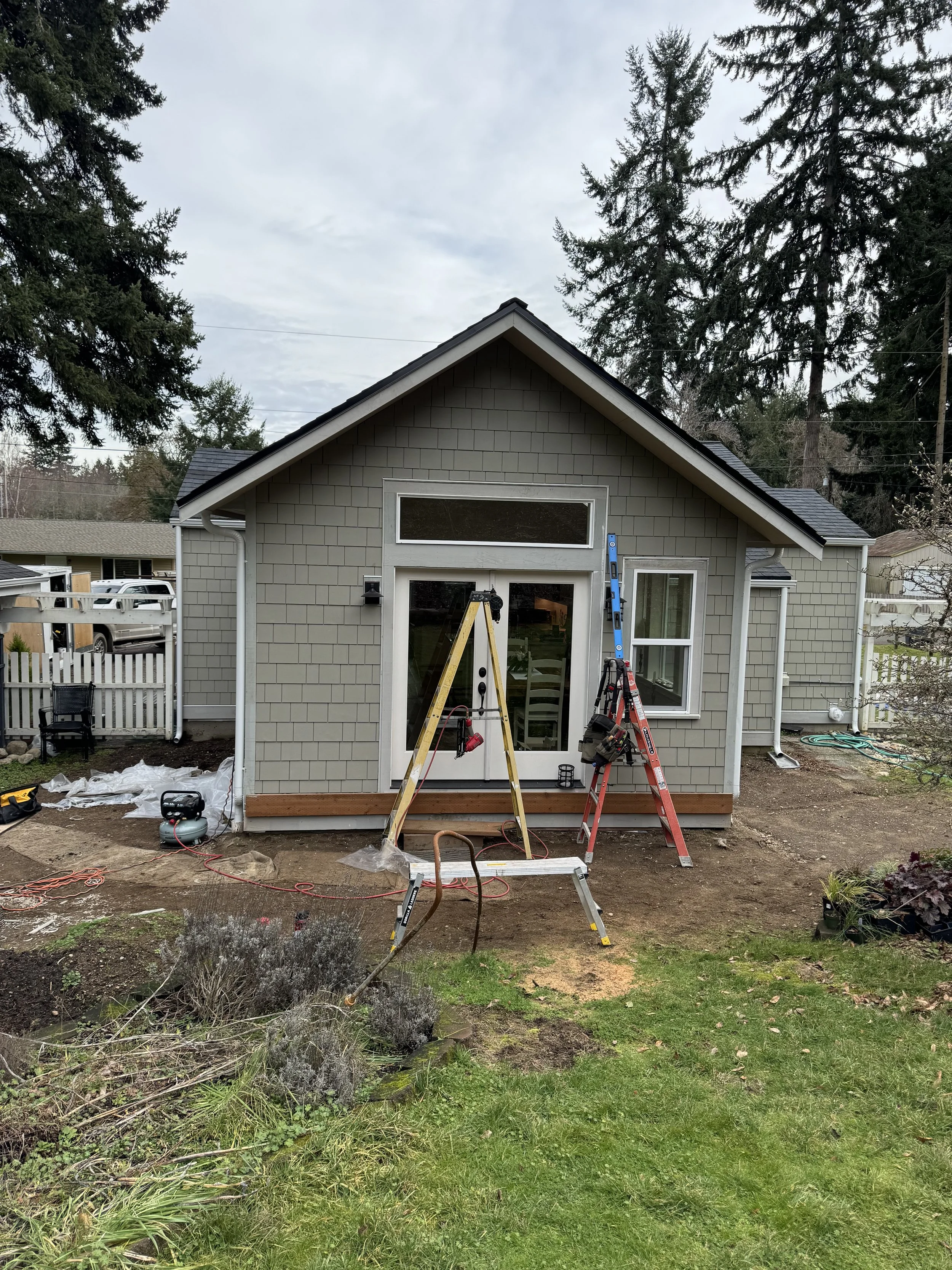 A house undergoing renovation with tools and ladders in front, plants and trees in the yard, and cloudy sky overhead.