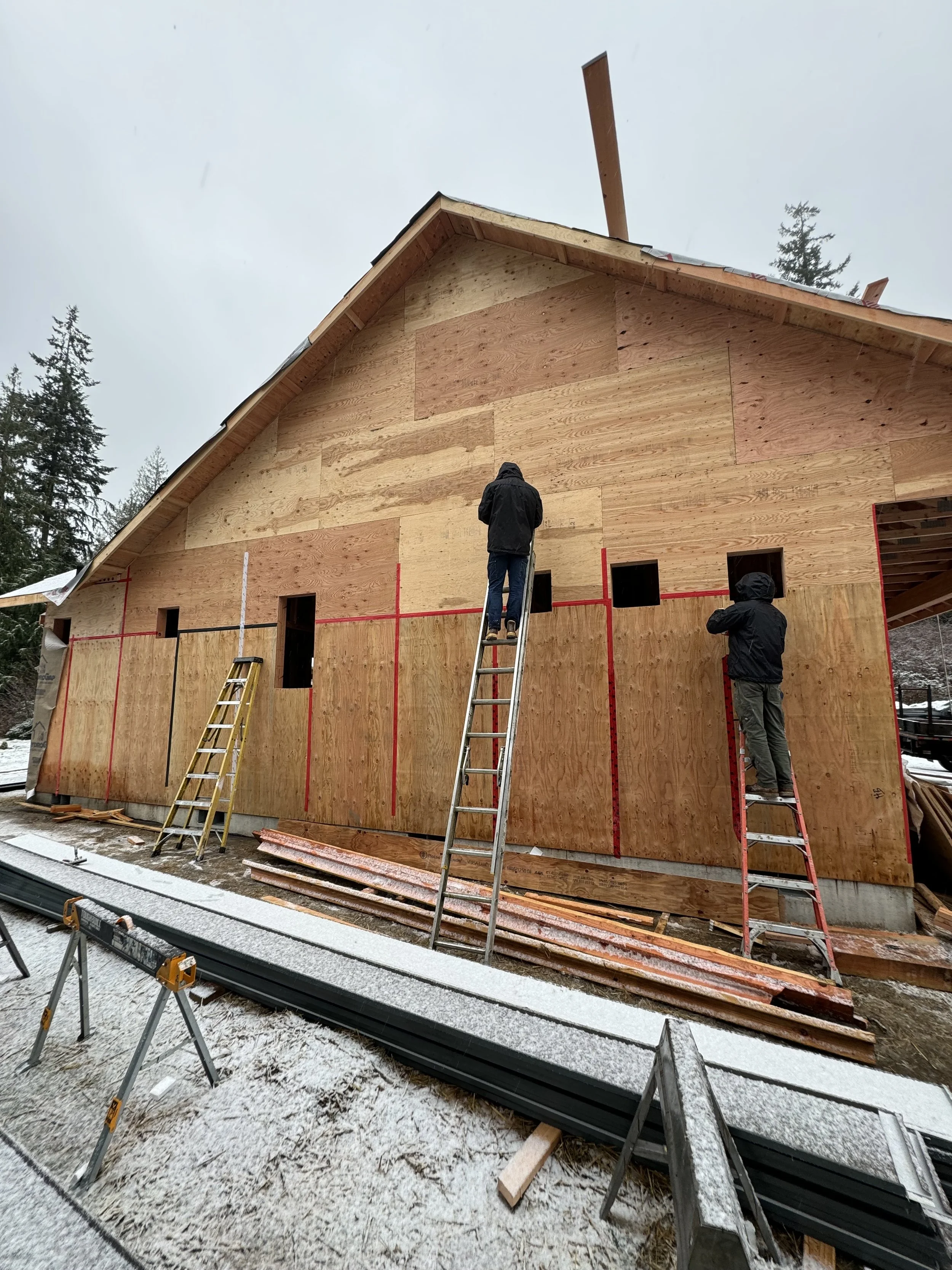 Workers constructing a wooden house, with ladders and construction materials around, on a snowy day.