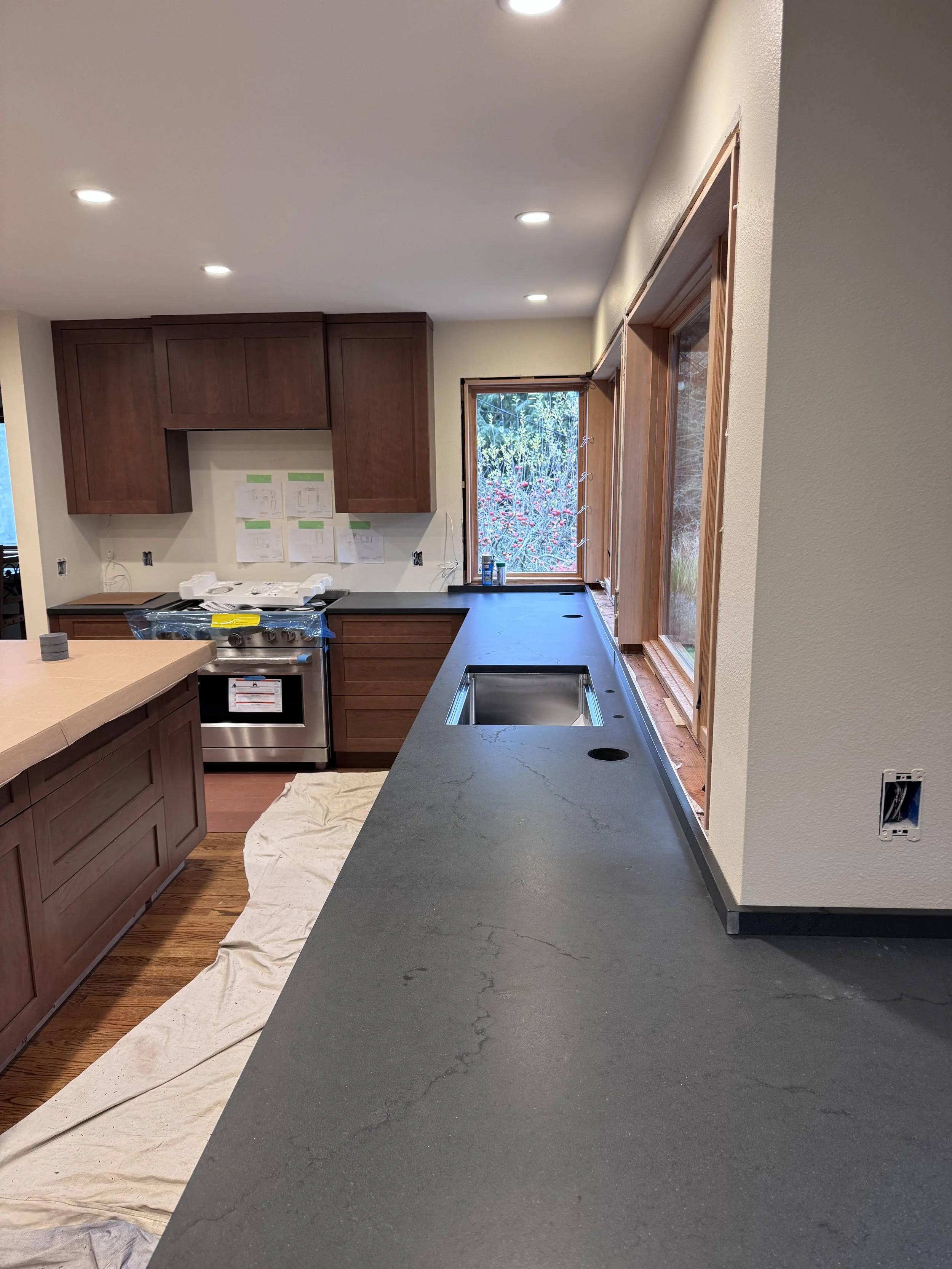 Kitchen under renovation with dark countertop, wooden cabinets, window showing a garden view