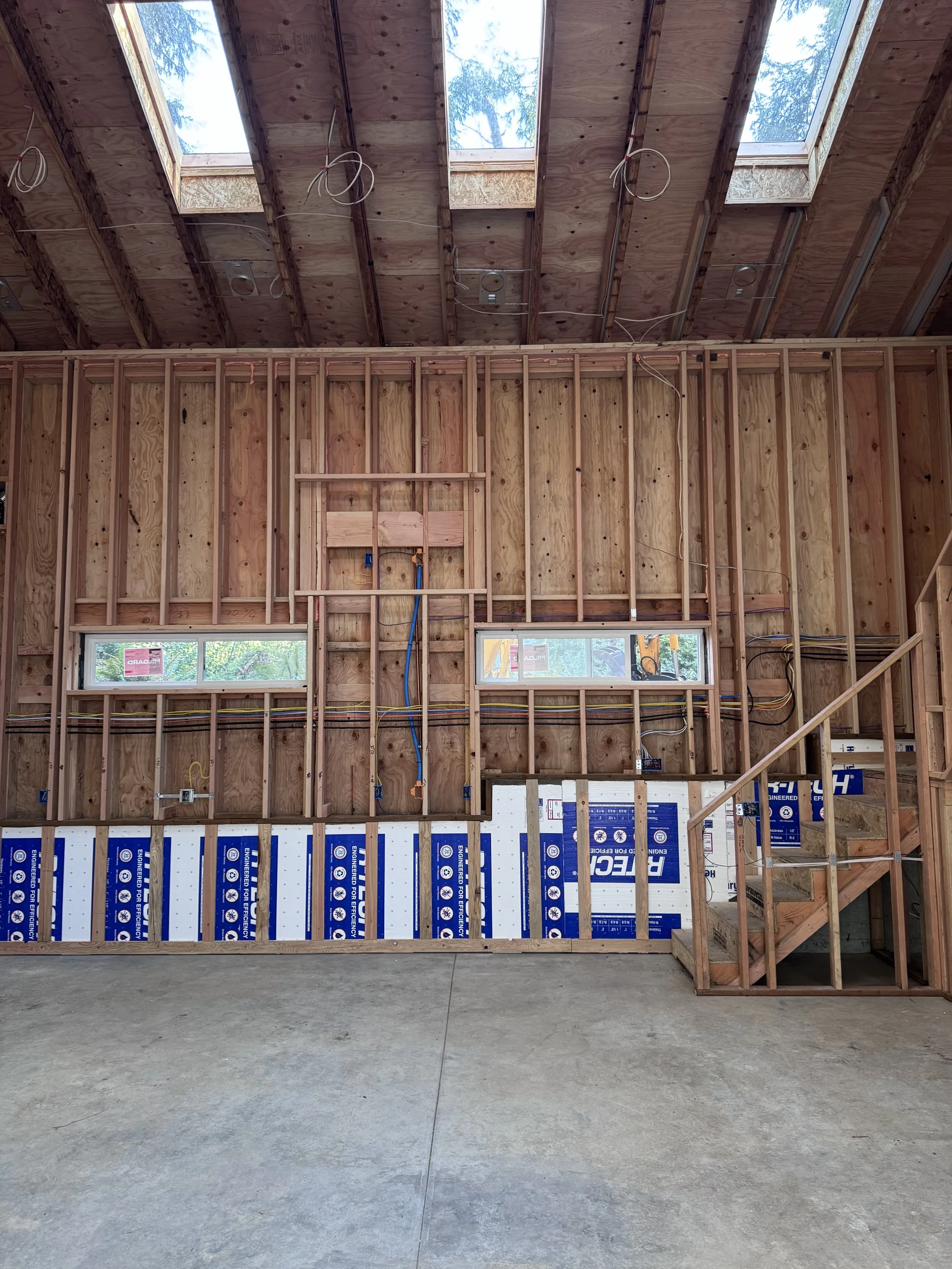 Interior of a house under construction with exposed wooden framing, partial walls, and a vaulted ceiling with skylights.
