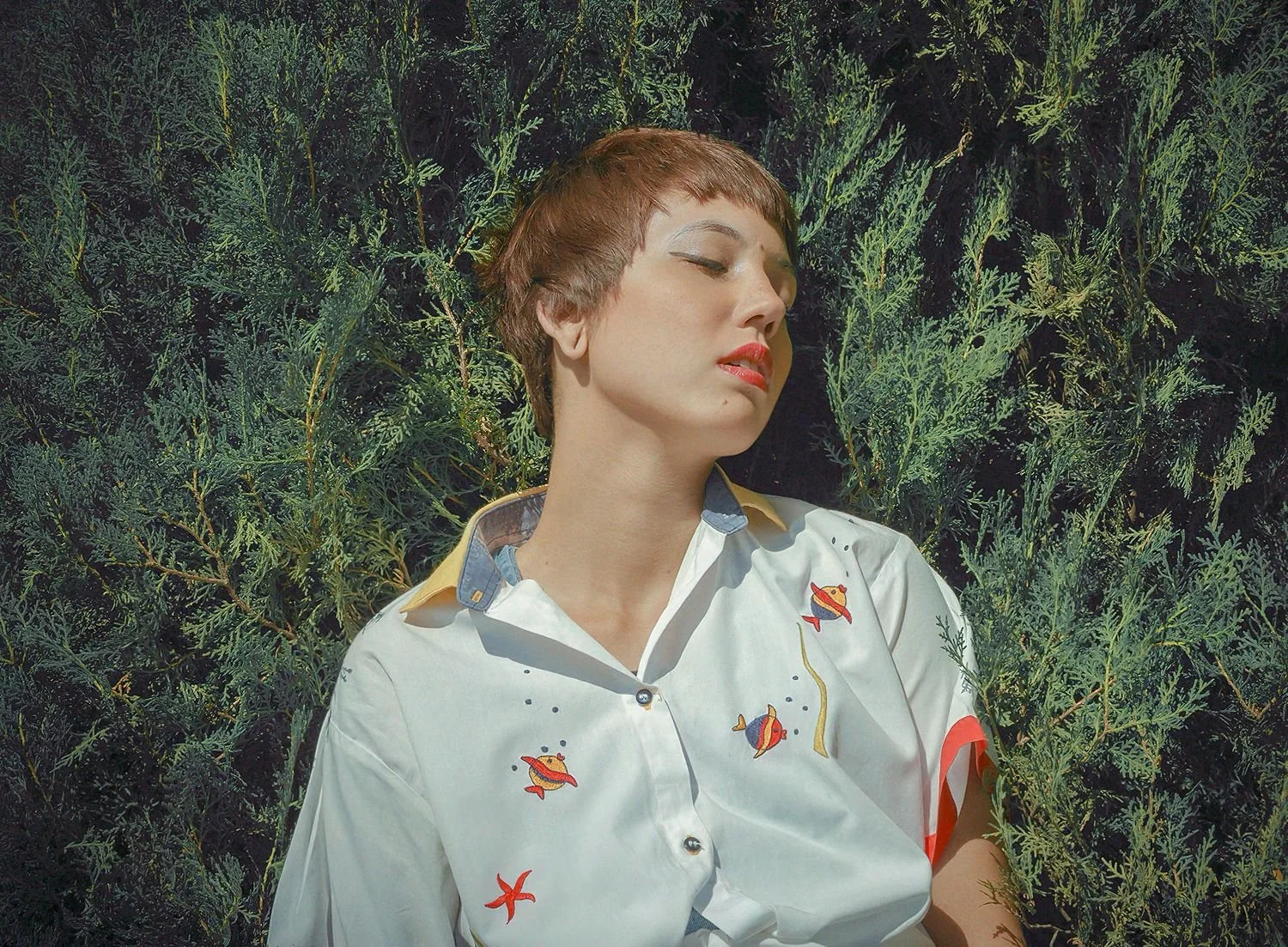 A woman with short brown hair and red lipstick, wearing a white shirt with aquatic-themed embroidery, is leaning against a dark green leafy background with her eyes closed.