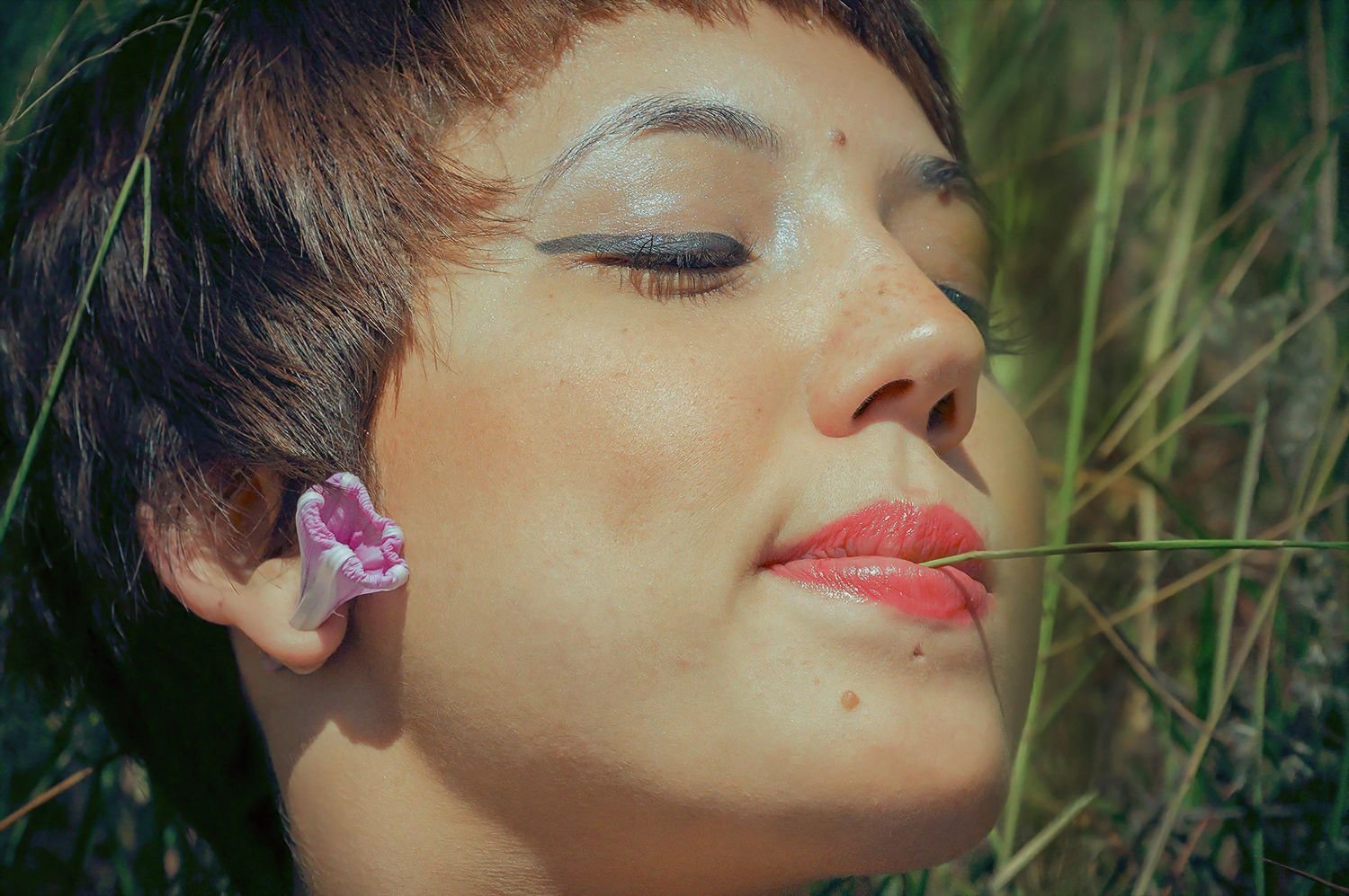 Close-up of a woman with short brown hair and makeup, lying among tall grass with her eyes closed, a blade of grass near her lips.