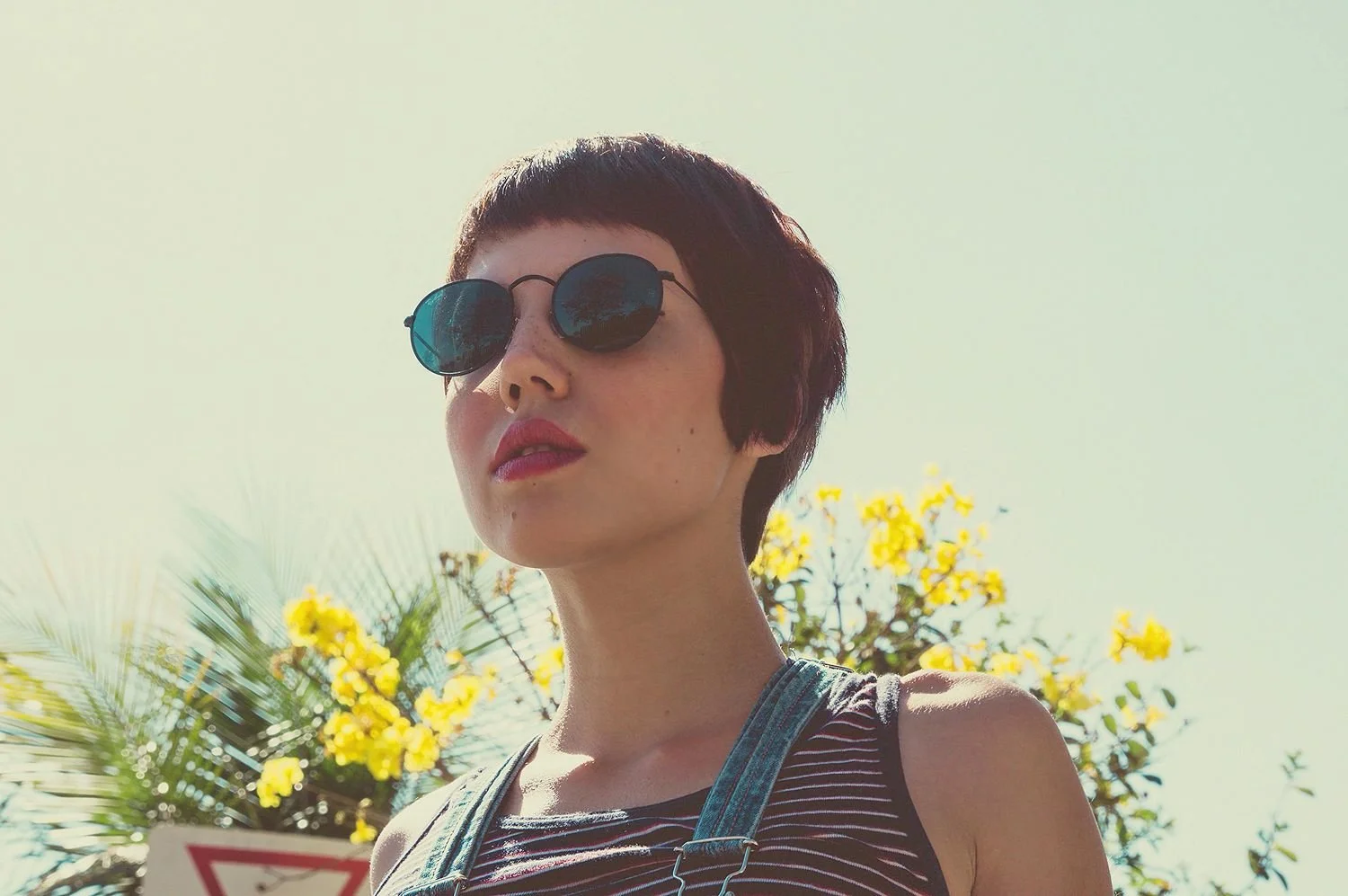 A woman with short dark hair and sunglasses looking up outdoors with yellow flowers and a clear sky in the background.