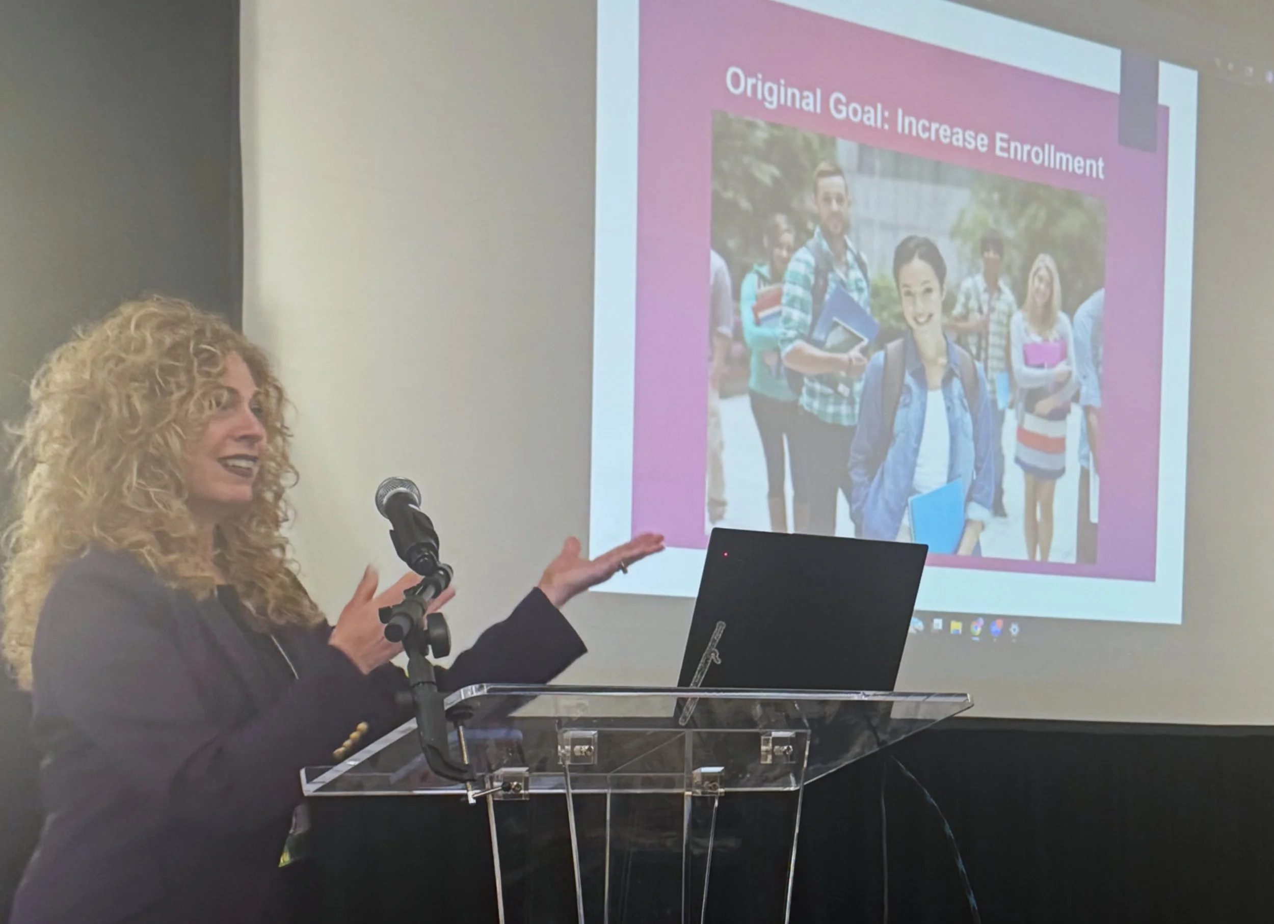 A woman with curly blonde hair giving a presentation at a podium with a microphone. A laptop is placed on the podium. A slide projected on a screen behind her shows a group of students holding books and backpacks and text that reads 'Original Goal: Increase Enrollment'.
