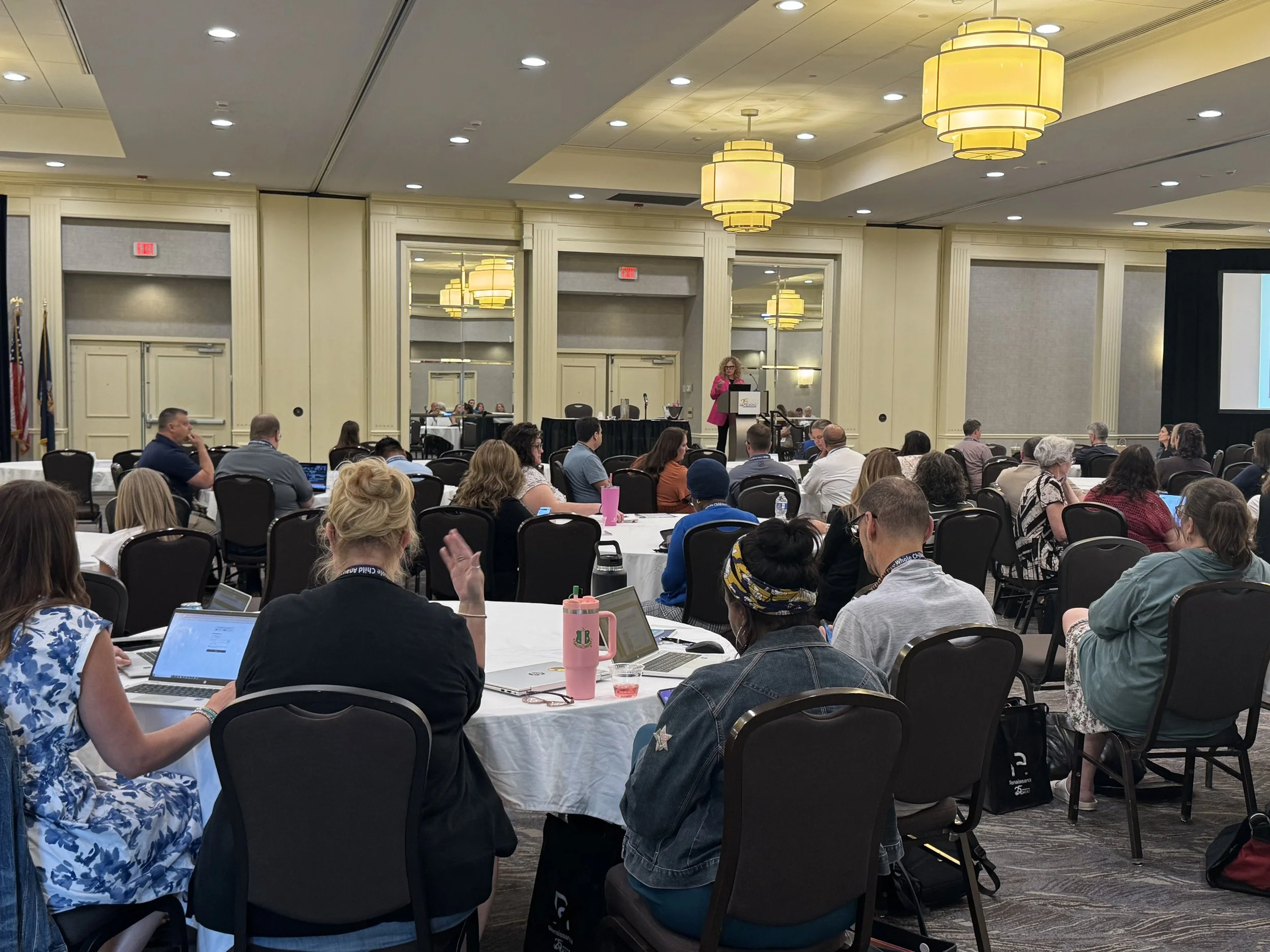 A large conference room filled with people sitting at round tables, listening to a woman speaking at a podium. The room has cream-colored walls, large mirrors, elegant light fixtures, and multiple overhead lights.