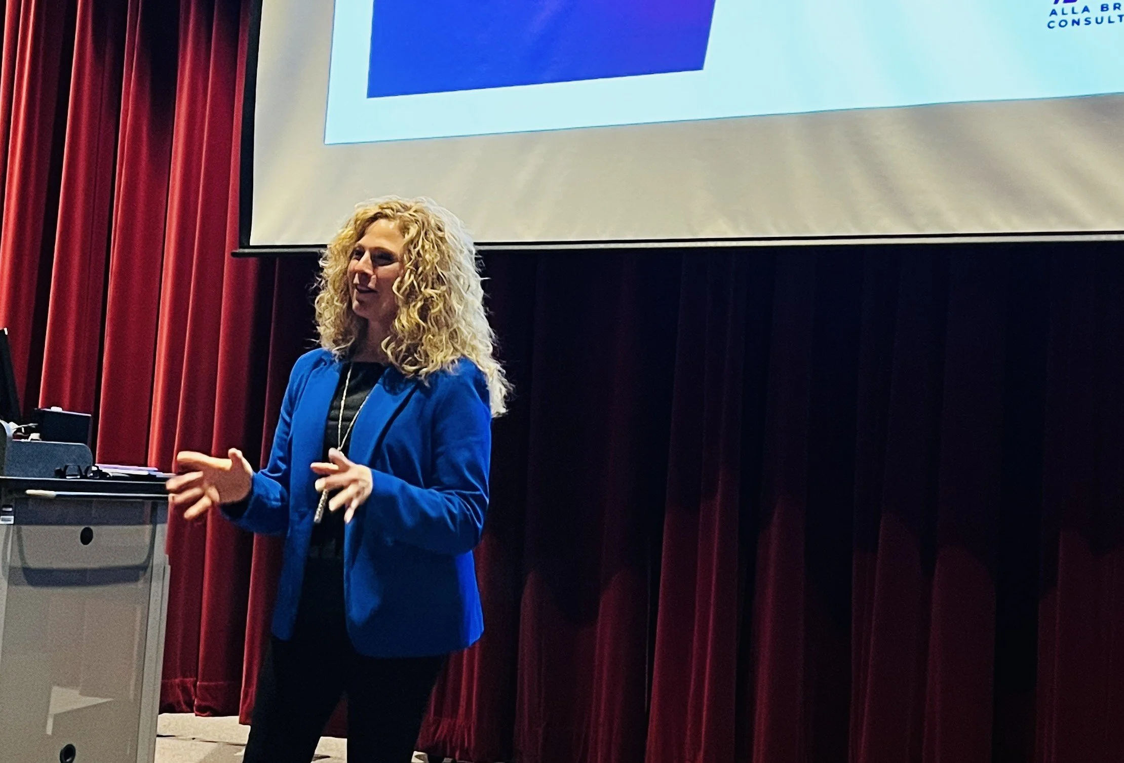 A woman with curly blonde hair wearing a blue blazer and black clothing, speaking on stage in front of a large screen with a presentation, with red curtains in the background.