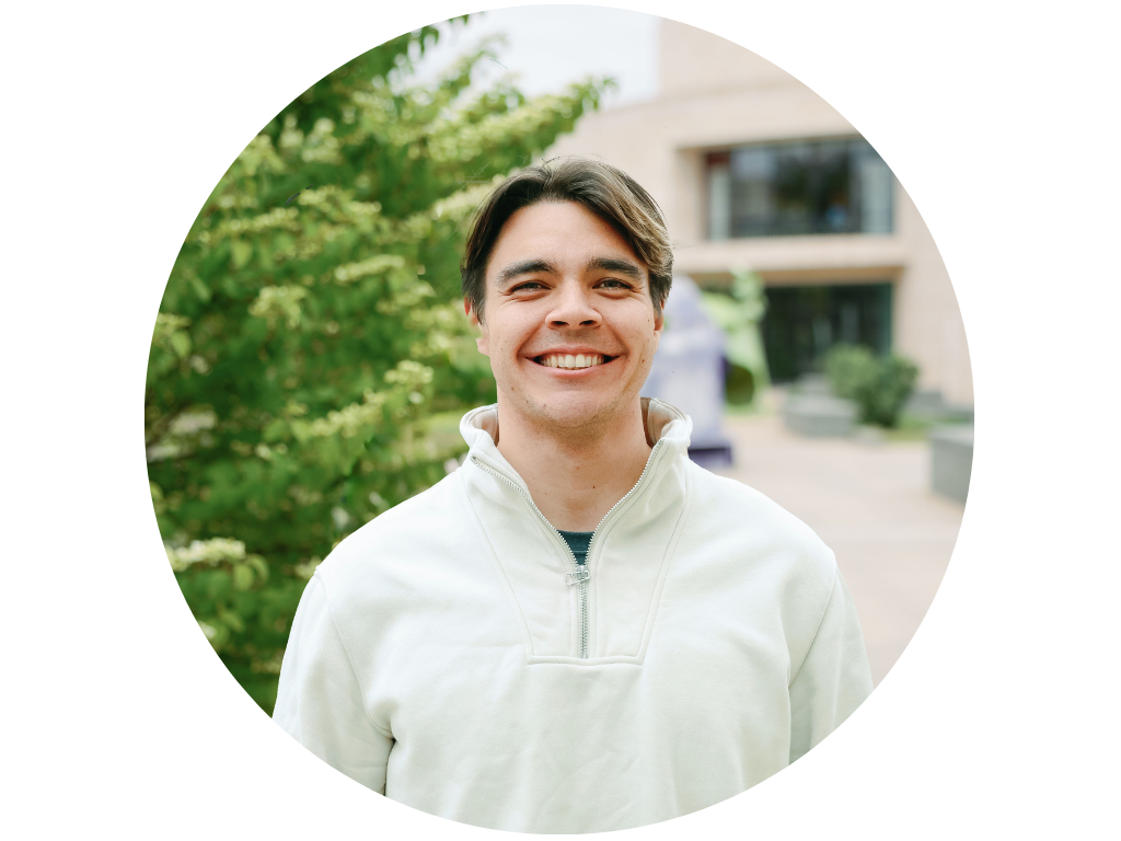 Smiling person in a gray button-up shirt, standing outdoors with trees and greenery in the background.