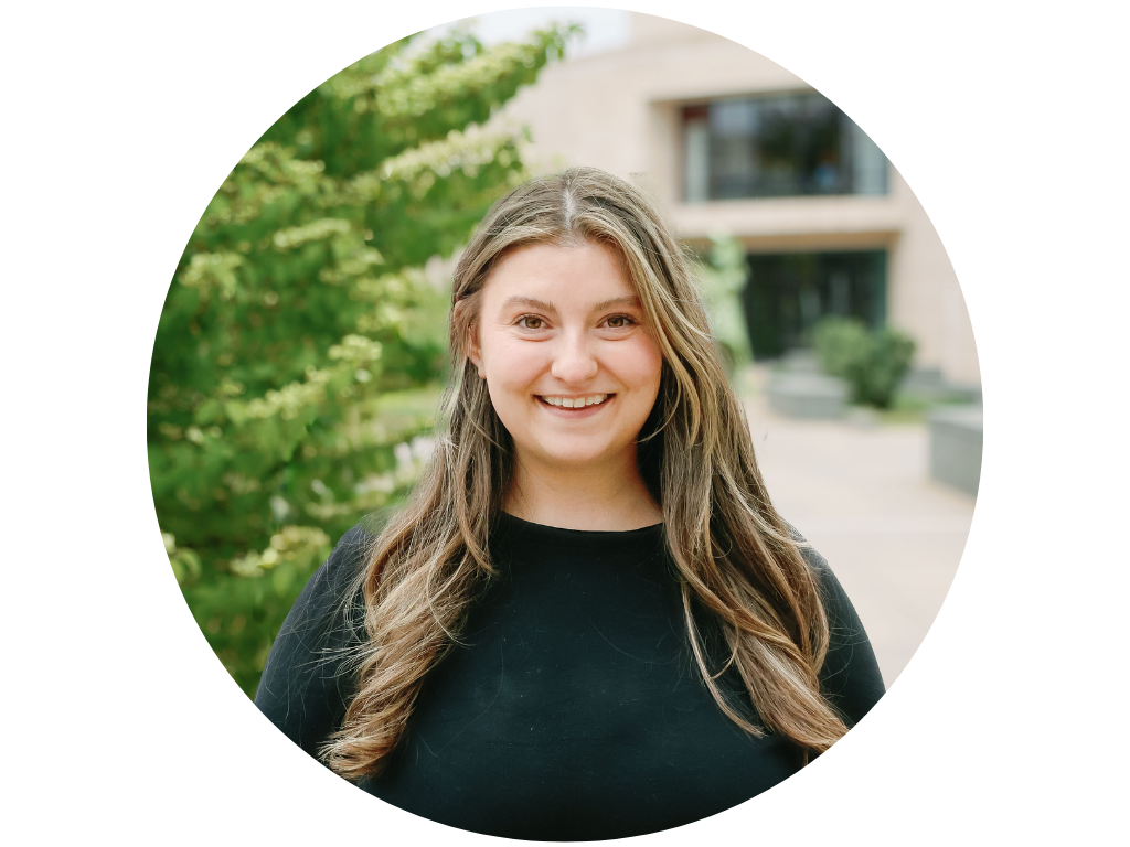 A woman with long wavy hair and a denim jacket smiling in a park setting with greenery and a lamp post in the background.