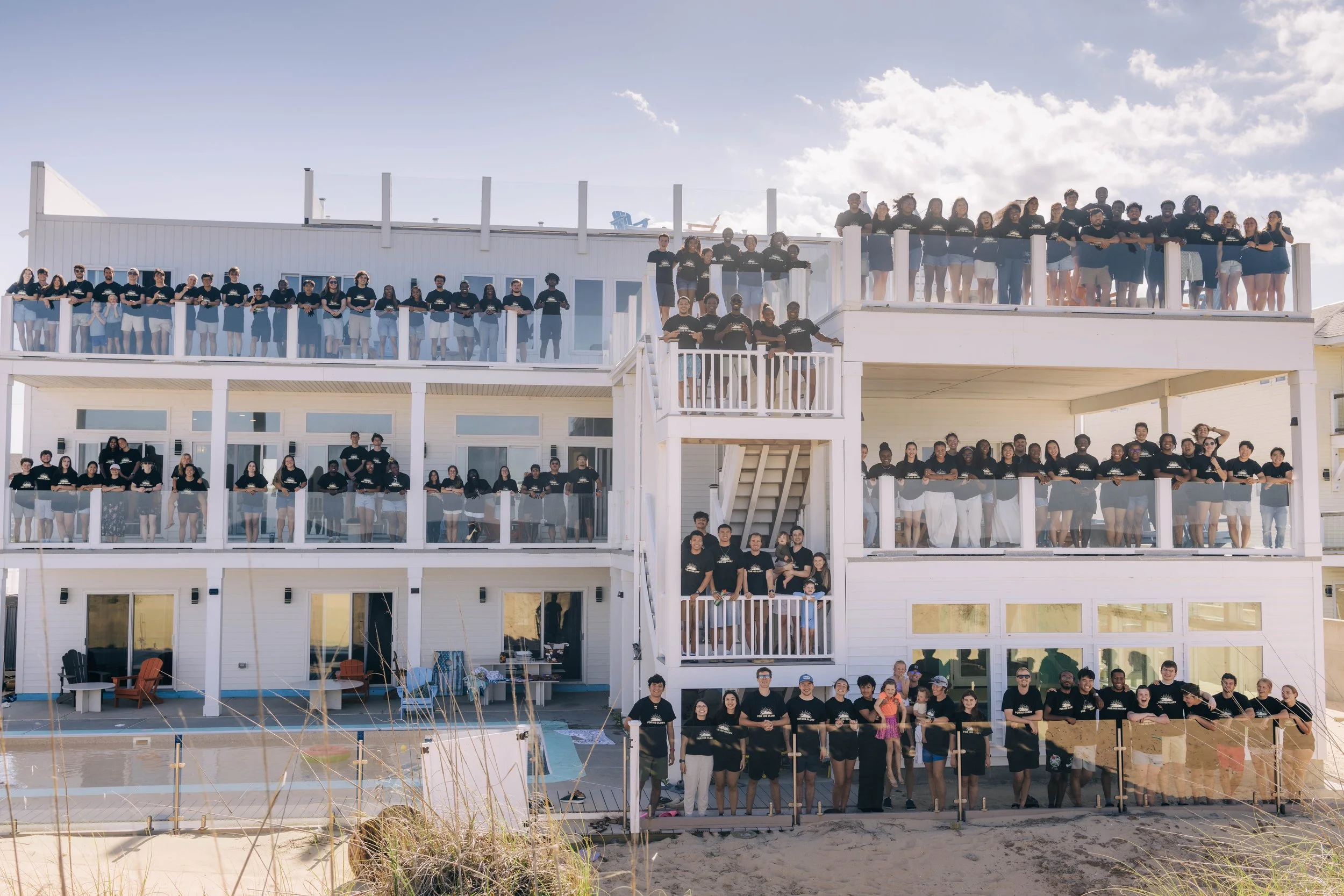 Large group of people on a sandy beach with a house in the background, all smiling and wearing casual white shirts, under a blue sky with clouds.