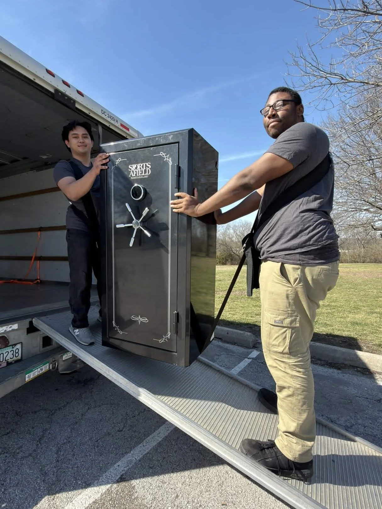 Two men are unloading a black safe with the logo 'Sports Afield' from a truck onto a metal ramp outdoors on a sunny day with clear blue skies. One man is standing on the truck, and the other is on the ground.