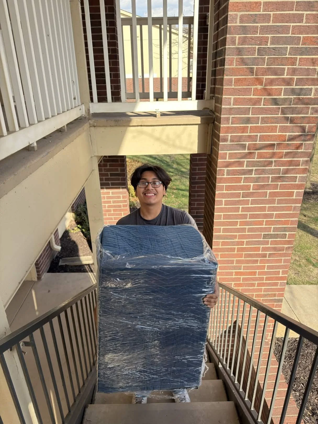 Young man with glasses smiling, holding a large wrapped object on a stairway outside of a brick apartment building.